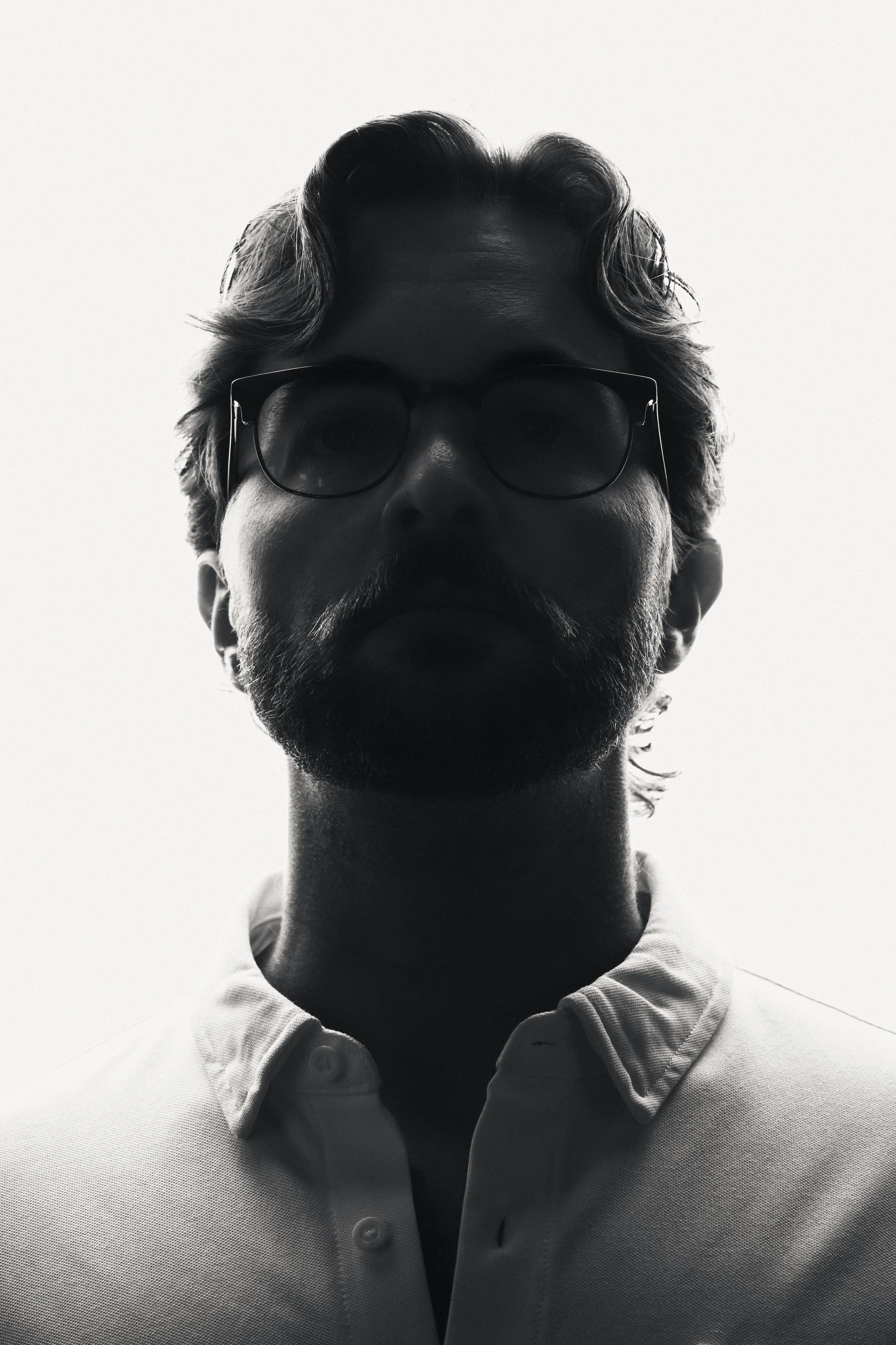 Backlit portrait of a man in a white button-up shirt, photographed from the shoulders up with strong rim light creating a silhouetted profile against a bright background, his facial features obscured.