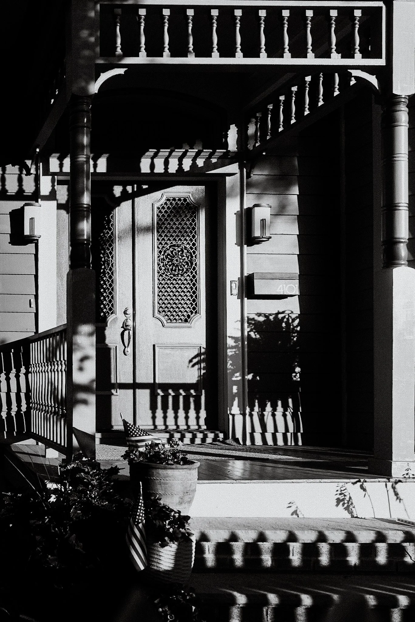 Black and white photo of the front porch of a house, featuring a door with ornate design, a staircase with rounded columns, and a porch railing. Potted plants with leafy foliage sit on the porch, and shadows cast by the sunlight create patterns on th