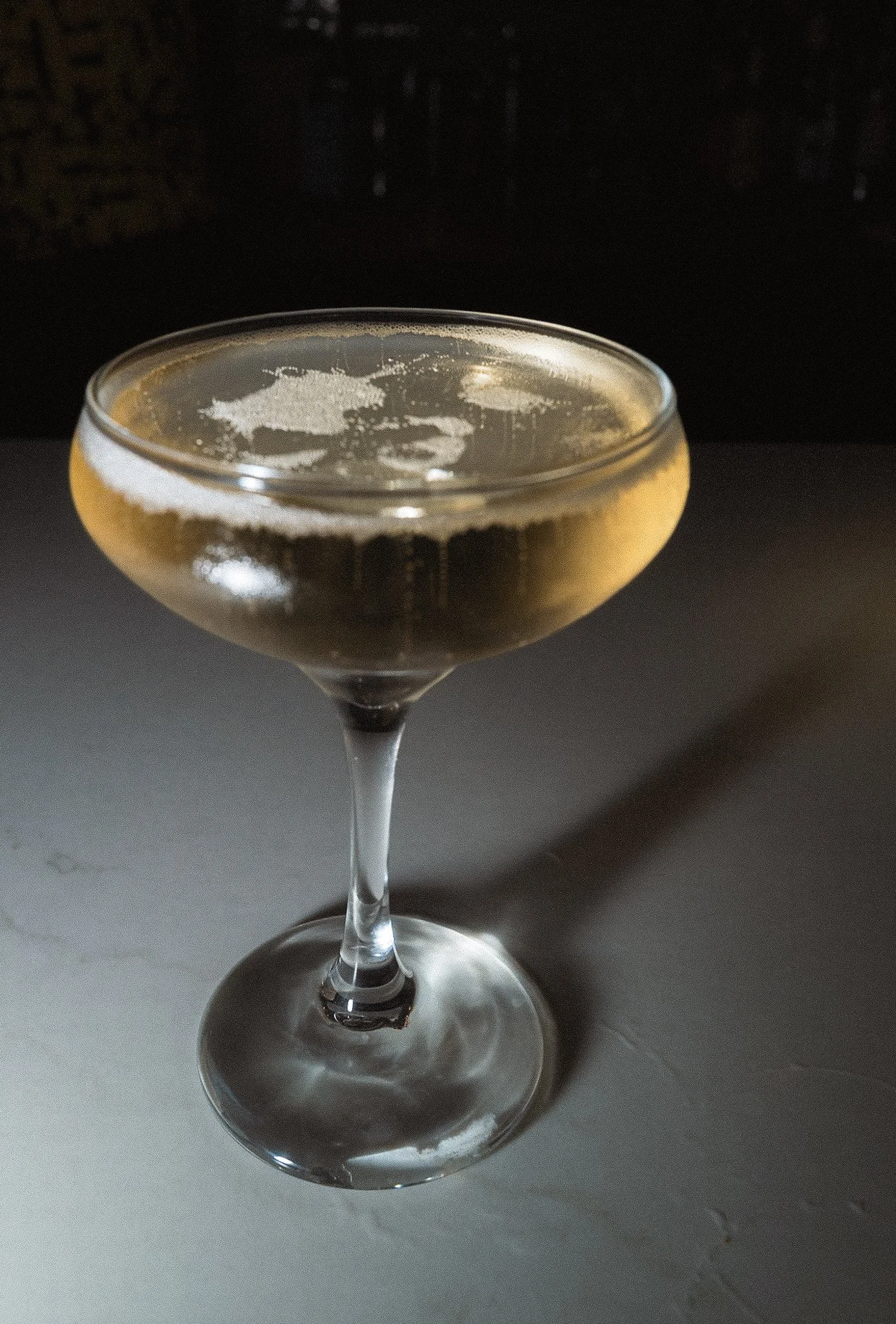 A cocktail glass filled with a light-colored beverage, topped with foam, placed on a surface with a dark background.