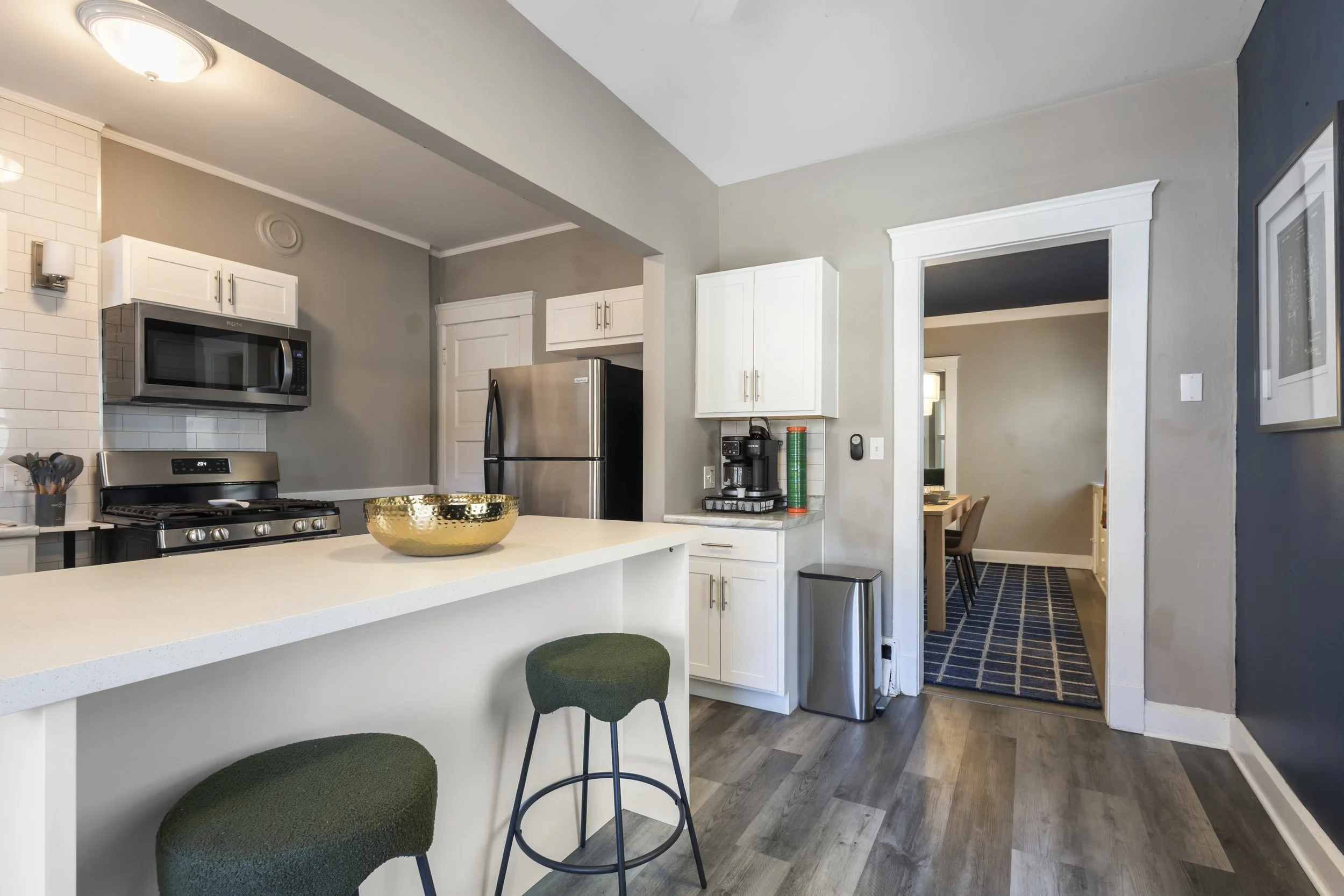A modern kitchen featuring white cabinetry, stainless steel appliances including a microwave and a refrigerator, a white countertop with a decorative gold bowl, and a small green cushioned stool. There is a doorway leading to a dining area with a tab
