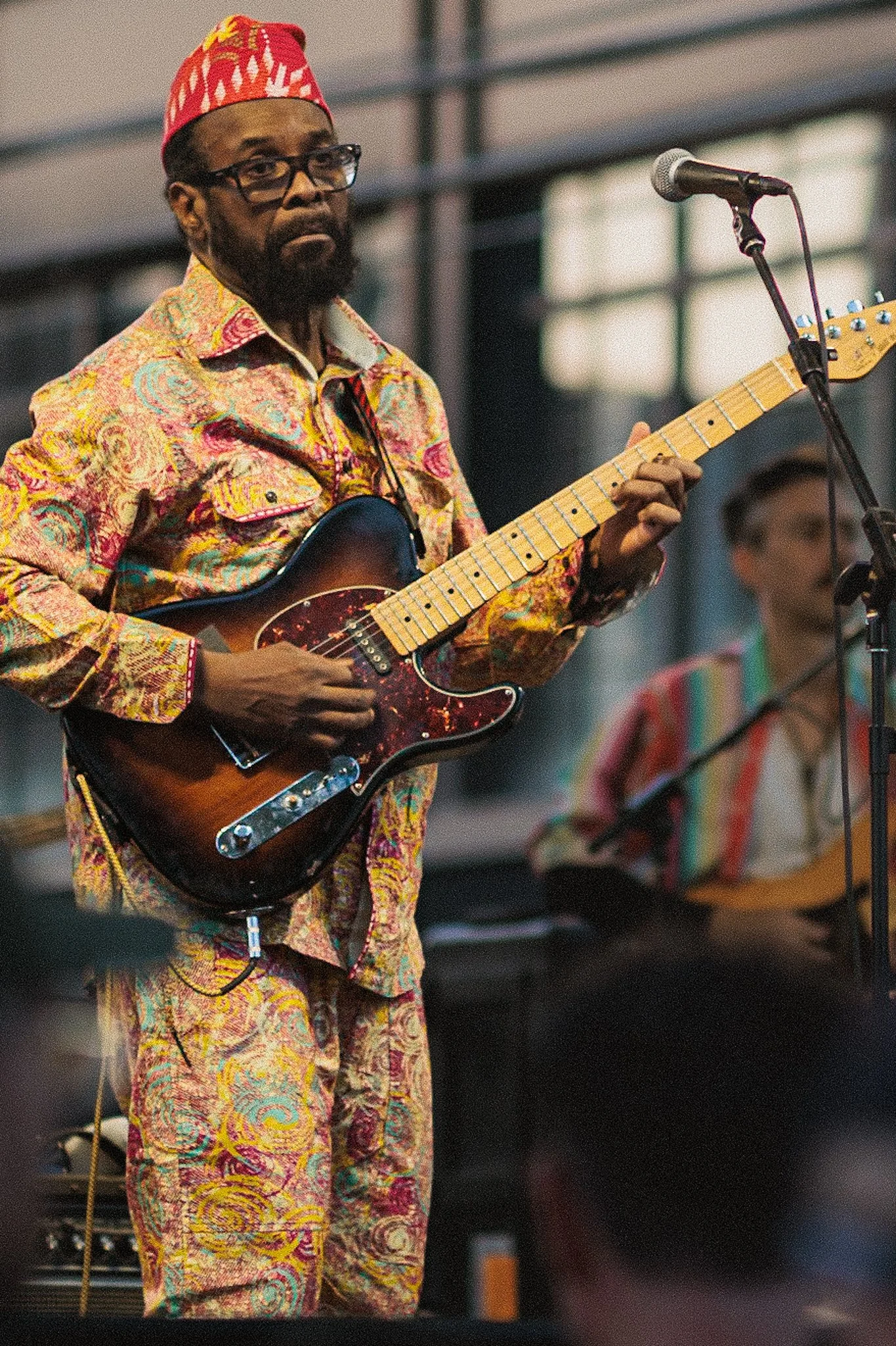 A man wearing colorful, paisley-patterned clothing and a red patterned cap playing an electric guitar on stage, with a microphone in front of him and another musician in the background.