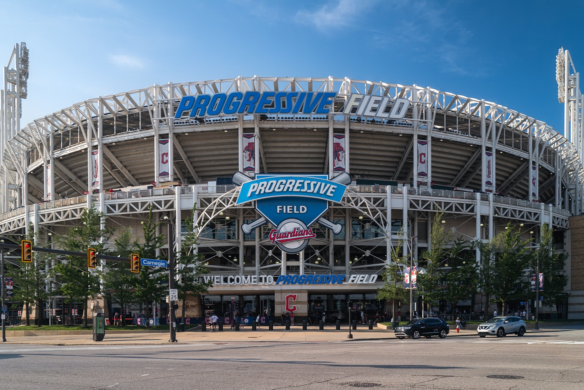 The exterior of Progressive Field, a baseball stadium, with large blue and gray signage and banners displaying the Cleveland Guardians logo. There are trees, traffic lights, and several cars in front of the stadium, along with pedestrians walking nea