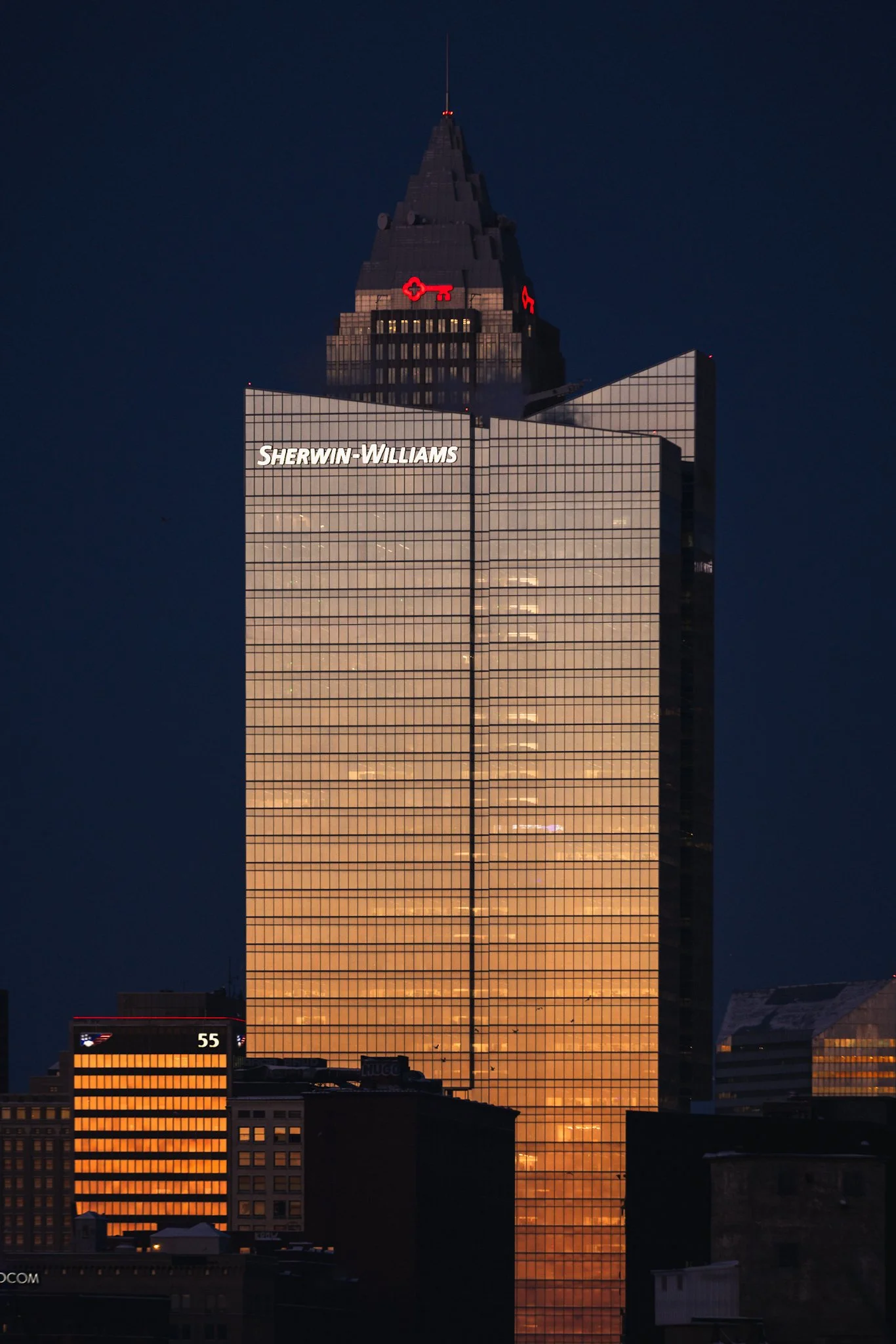 The Sherwin-Williams Global Headquarters, a sleek 36-story glass tower in downtown Cleveland, Ohio, glows in warm golden hour sunlight. Its distinctive spire and illuminated logo shine against a deep blue hour sky at dusk, with surrounding buildings 