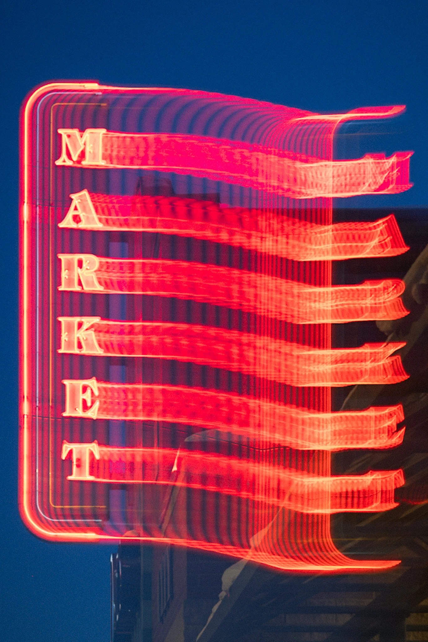 A dynamic long-exposure photograph captures a tall neon "MARKET" sign glowing in vibrant pinkish-red against a deep twilight blue sky. Multiple overlapping streaks and motion blur create a wavy, ethereal trail effect along the sign's white block lett