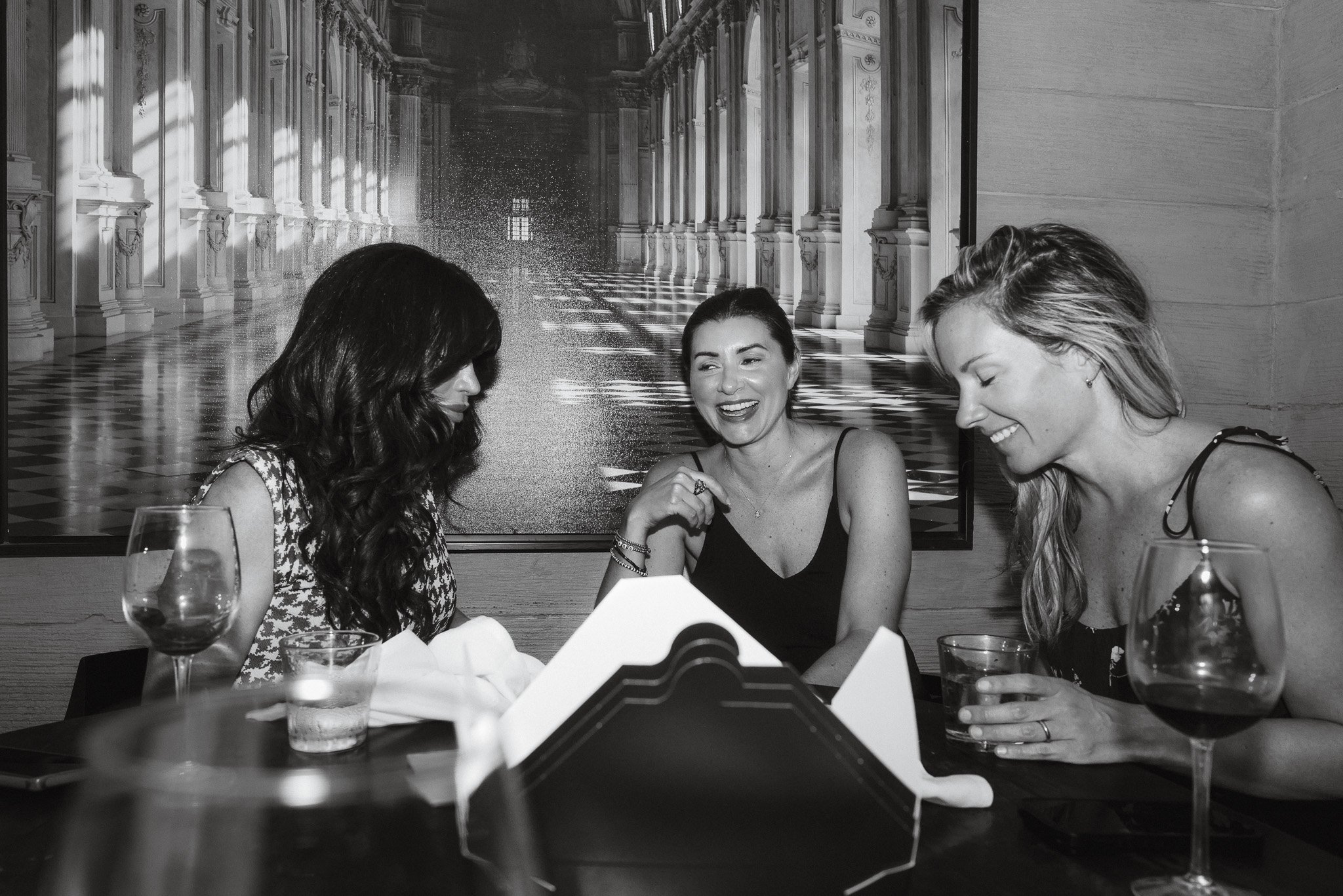 Three women sitting at a table in a restaurant, laughing and smiling. Glasses of water and wine are on the table, with a large decorative painting of a grand hall in the background.