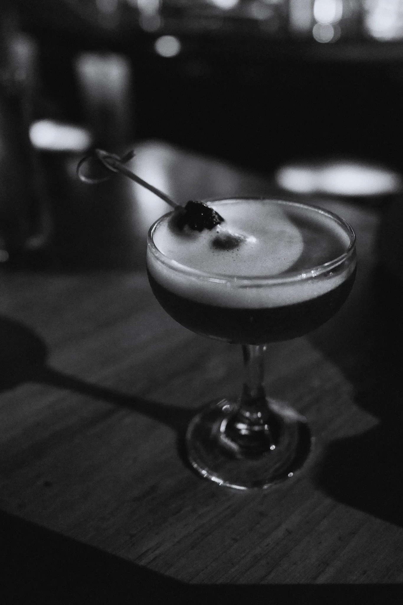 Black and white photo of a cocktail in a coupe glass with a cherry on top, placed on a wooden table.