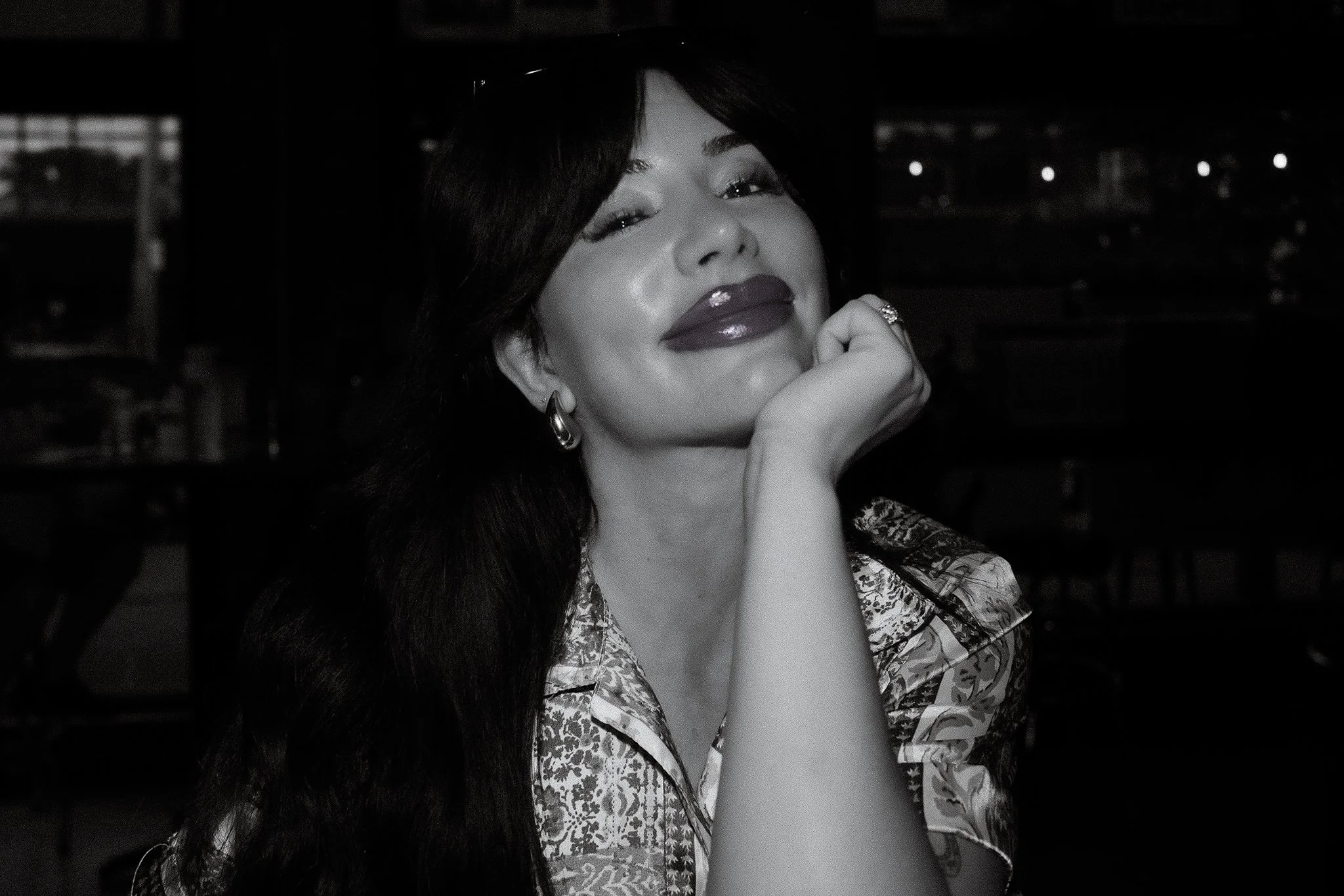 A woman with long dark hair, wearing makeup with shiny lips, resting her chin on her hand, in a dimly lit setting.