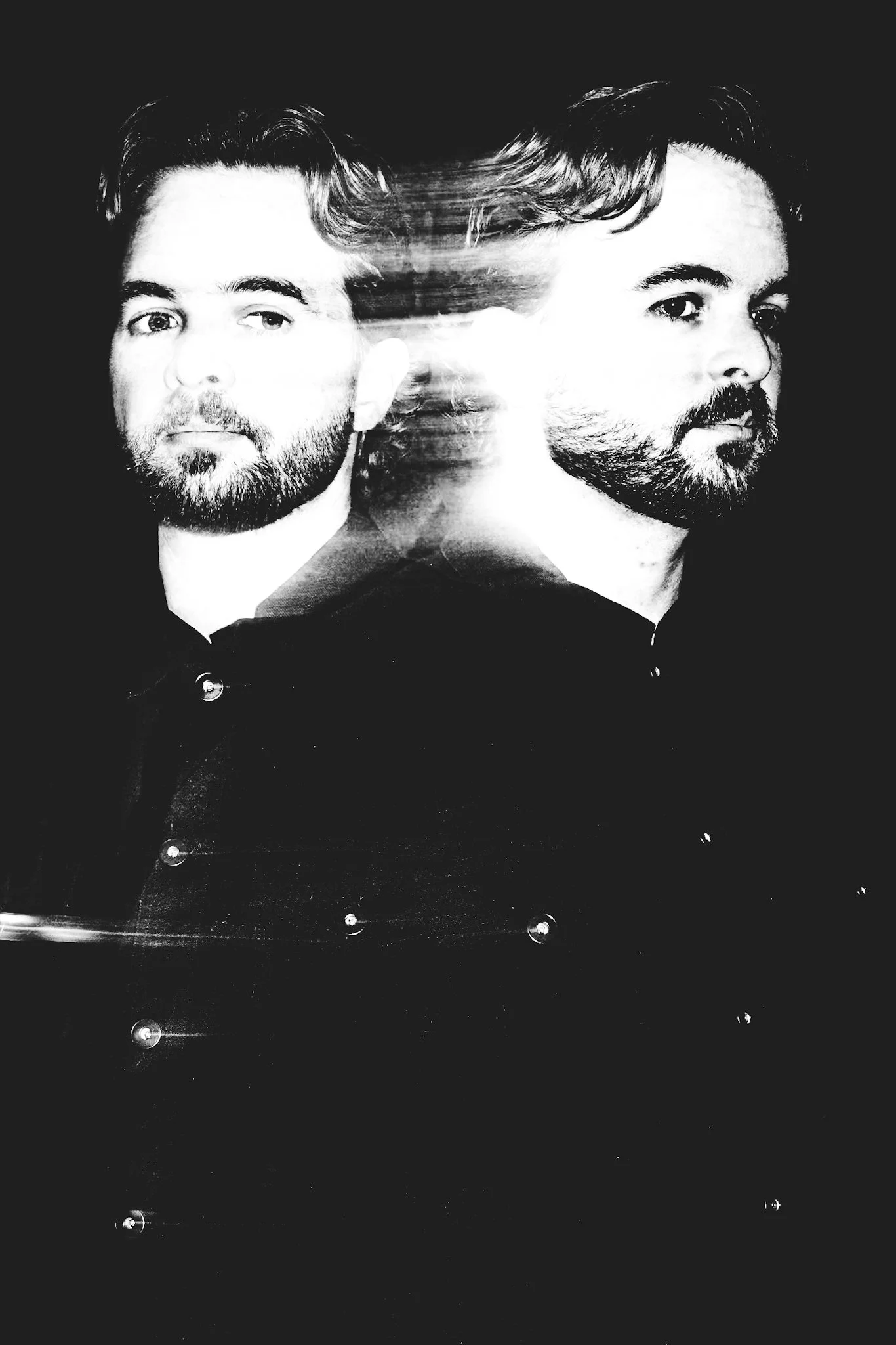 High-contrast black and white portrait with a mirrored, double-exposed figure against a dark background, the face blurred by motion and multiple flashes while metallic buttons on the shirt create small points of light.