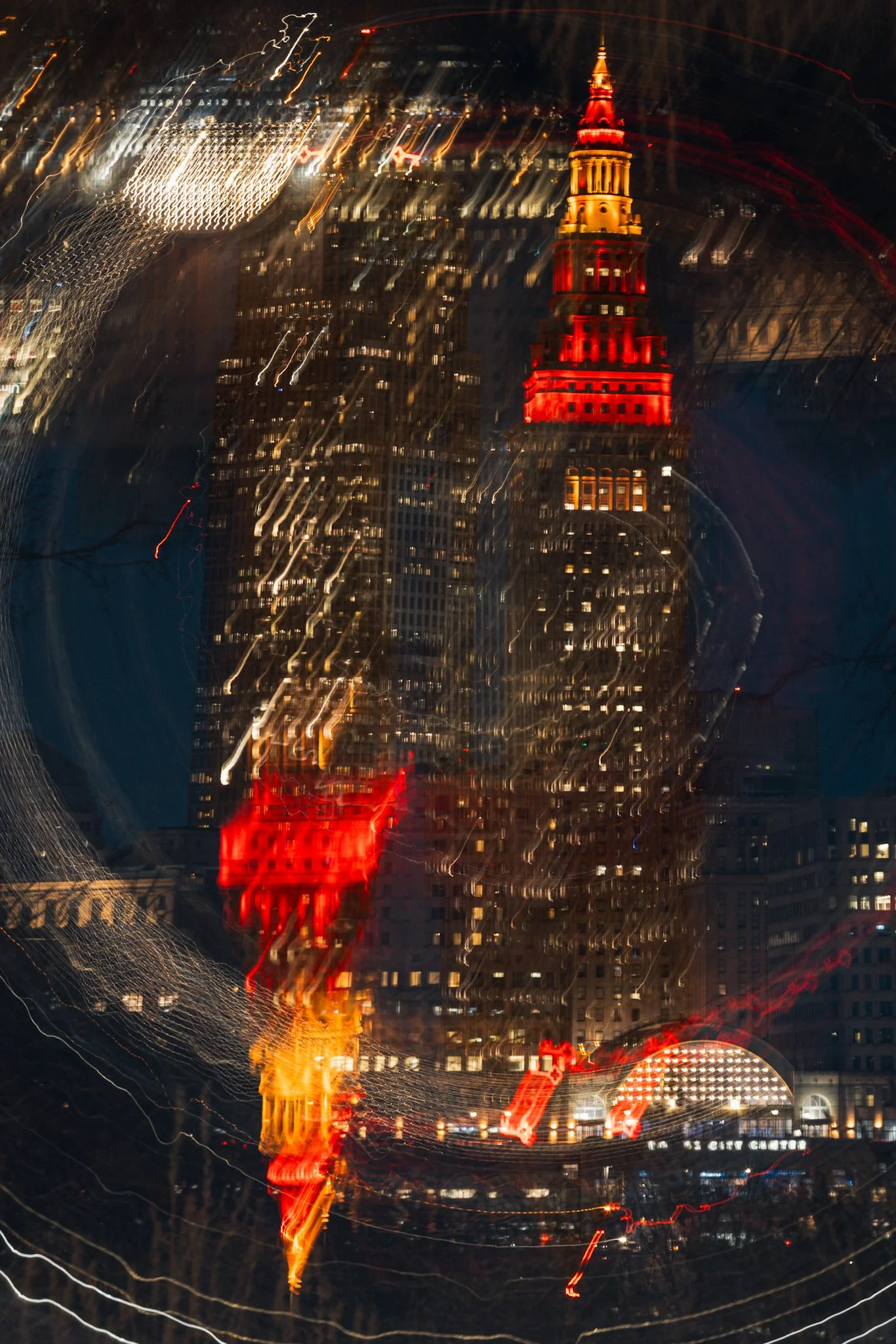 A nighttime photo of Cleveland’s Terminal Tower lit up in red and gold, with abstract swirling light trails creating a layered, motion-blur effect across the image.
