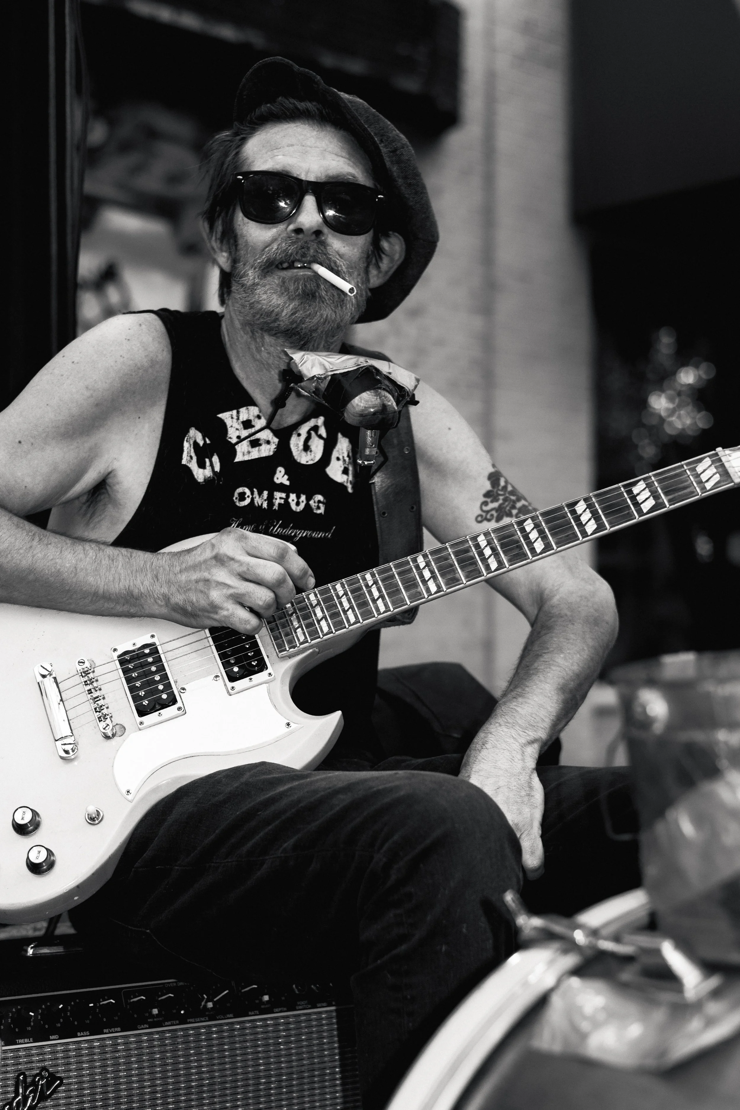 Black and white photo of a street musician sitting outdoors with an electric guitar on his lap. He wears sunglasses, a beret, and a sleeveless CBGB T-shirt, with a cigarette hanging from his mouth. A harmonica rests in a holder around his neck, and h