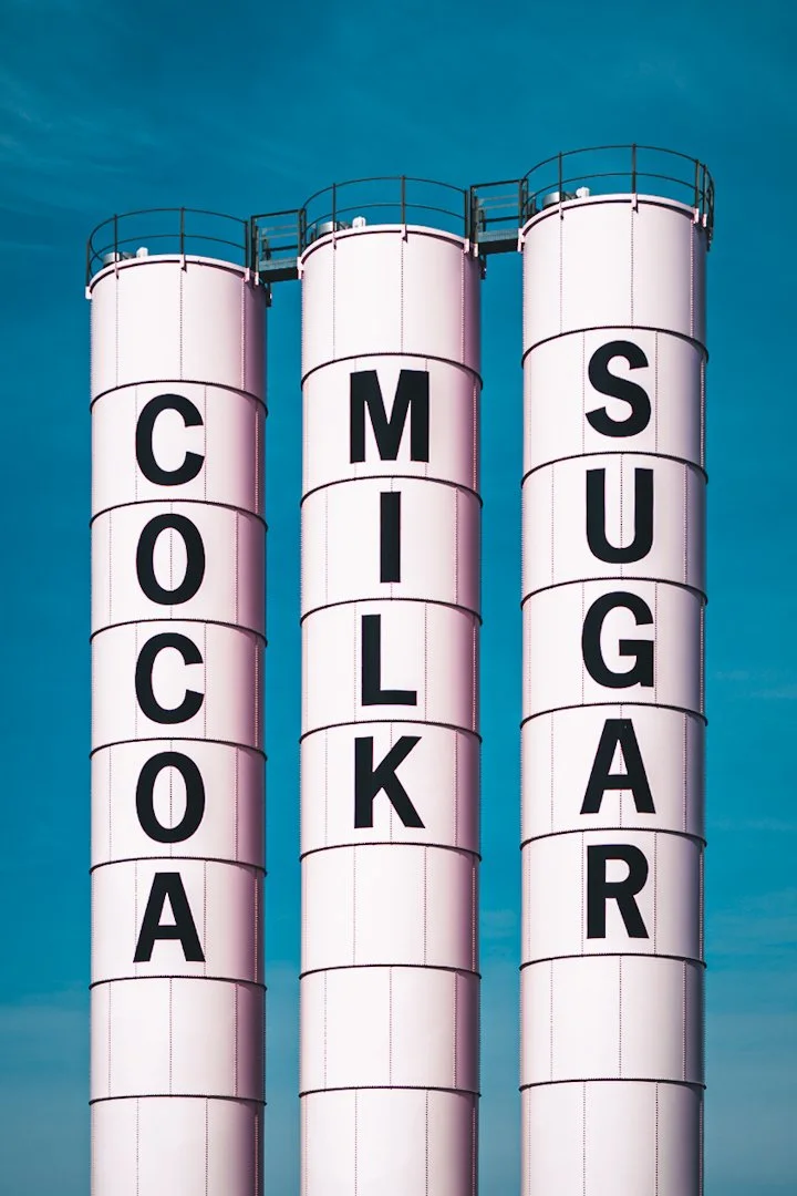 Three tall white silos at Malley's Chocolates in Cleveland, labeled "COCOA," "MILK," and "SUGAR" in bold black letters, standing symmetrically against a clear blue sky.