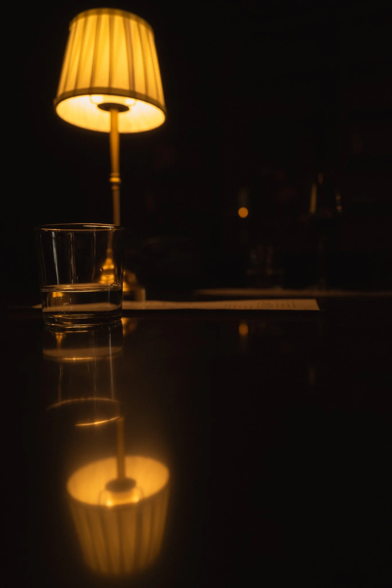 A table lamp with a beige lampshade and a yellow glow, positioned on a dark surface in a dimly lit room, with a glass of water reflecting the lamp's light.