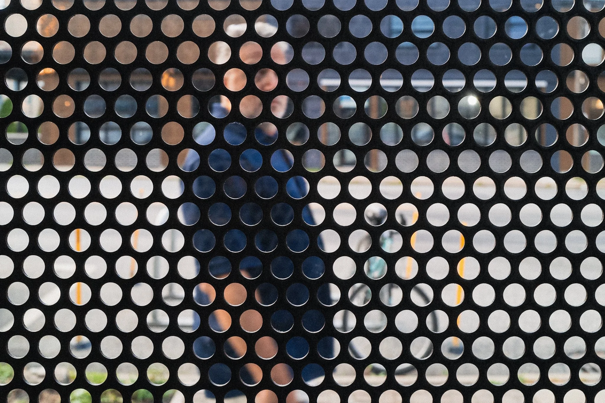 A black metal fence with circular holes through which two people can be seen in the background.