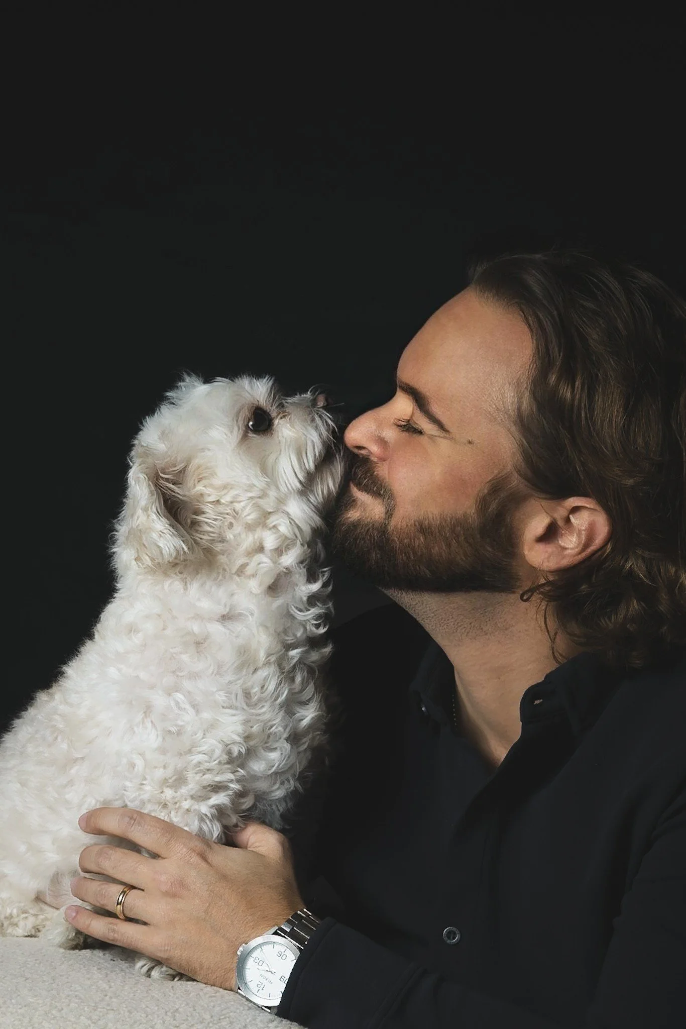 A man with long hair and a beard holding and kissing a small, curly-haired white dog against a black background.