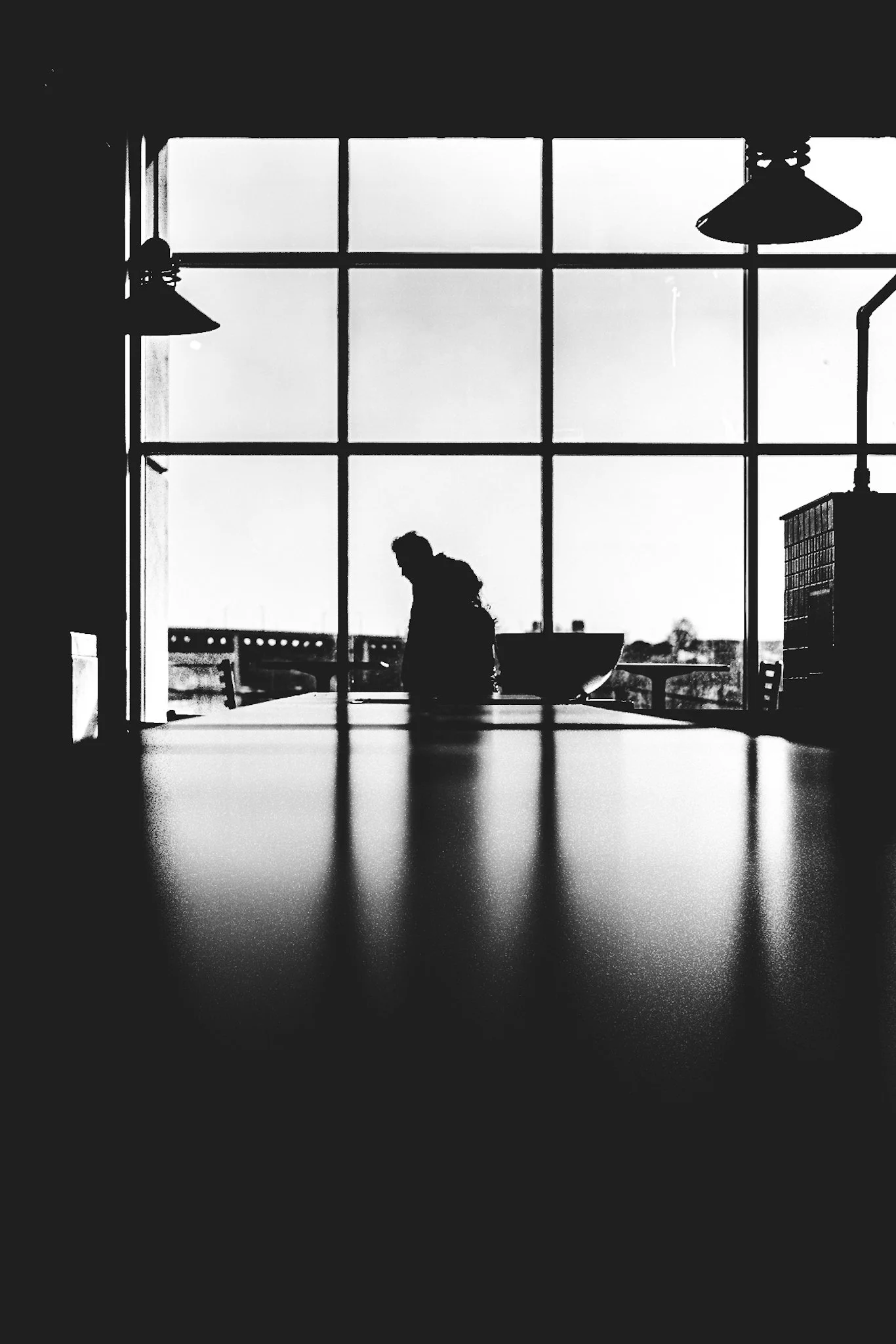 This black-and-white photograph captures a silhouetted figure of a man with a backpack, standing pensively at a long wooden table viewed from behind, framed by large industrial windows. Sunlight streams in, casting soft reflections on the glossy tabl