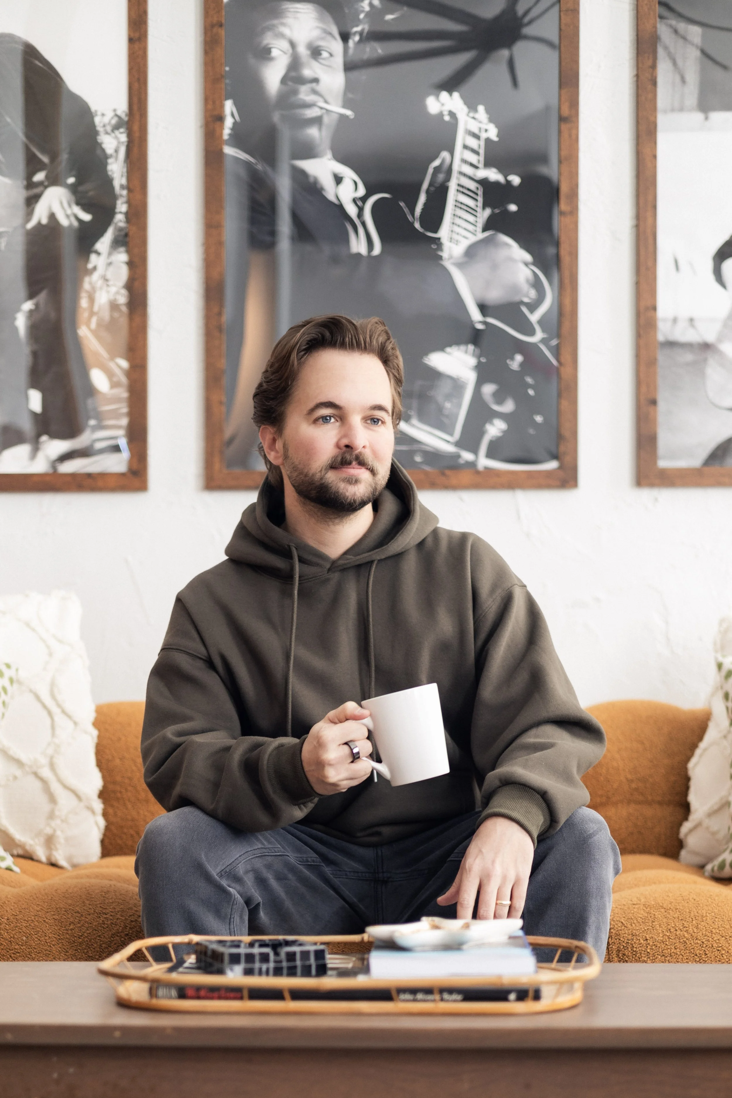 Matt Rutter sits on a mustard-colored couch in a cozy living room, wearing an moss hoodie and jeans, holding a white coffee mug, with framed picture of BB King and black-and-white guitar hanging on the wall behind him and a tray of books and small it