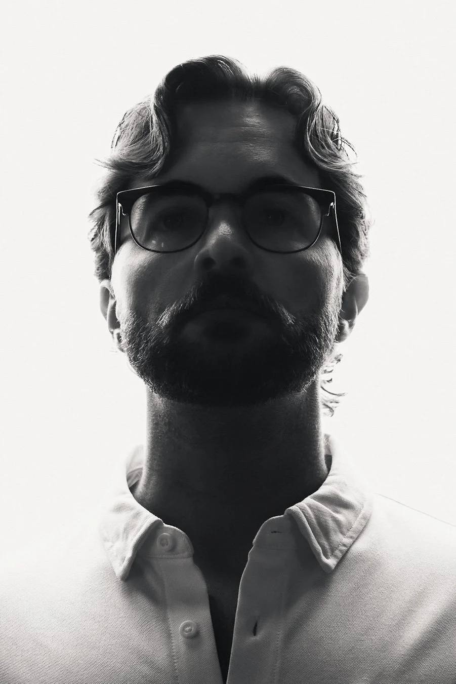 Backlit portrait of a man in a white button-up shirt, photographed from the shoulders up with strong rim light creating a silhouetted profile against a bright background, his facial features obscured.