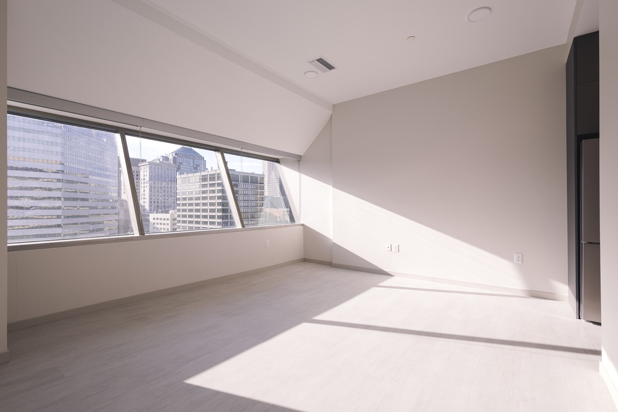 Spacious empty apartment room in Cleveland with light floors and large slanted windows filling the space with sunlight and a view of downtown office buildings.