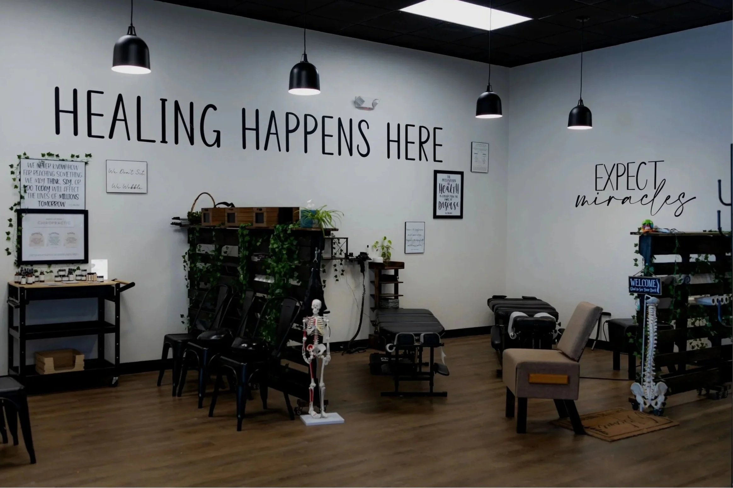 Interior of a holistic health or chiropractic clinic with the phrase 'Healing Happens Here' on the wall, various medical equipment, a skeleton model, and motivational signs.