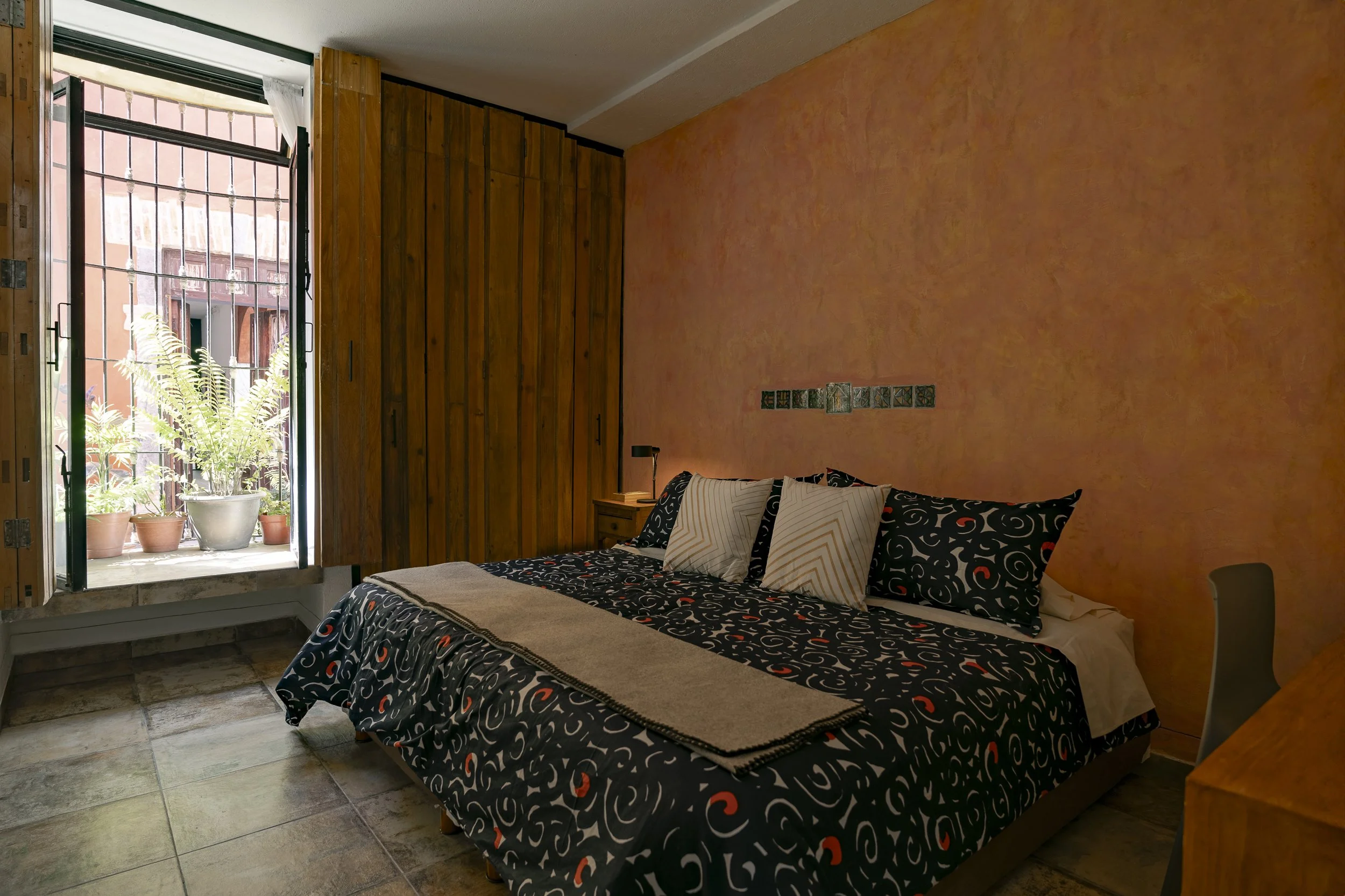 Habitación con cama, almohadas, pared color terracota y puerta abierta que da a un balcón con plantas