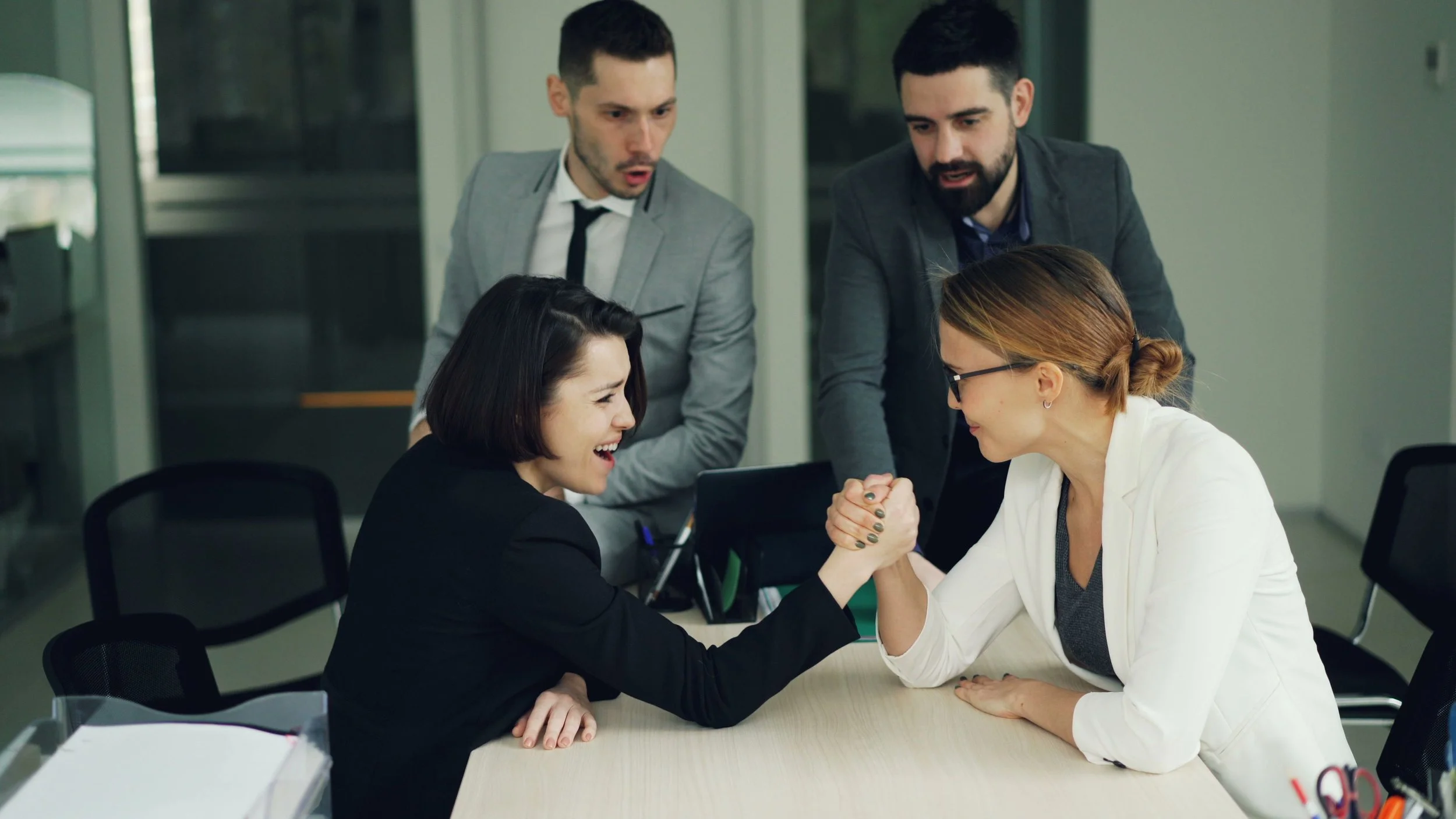 Two businesswomen arm wrestling at a conference table while colleagues watch