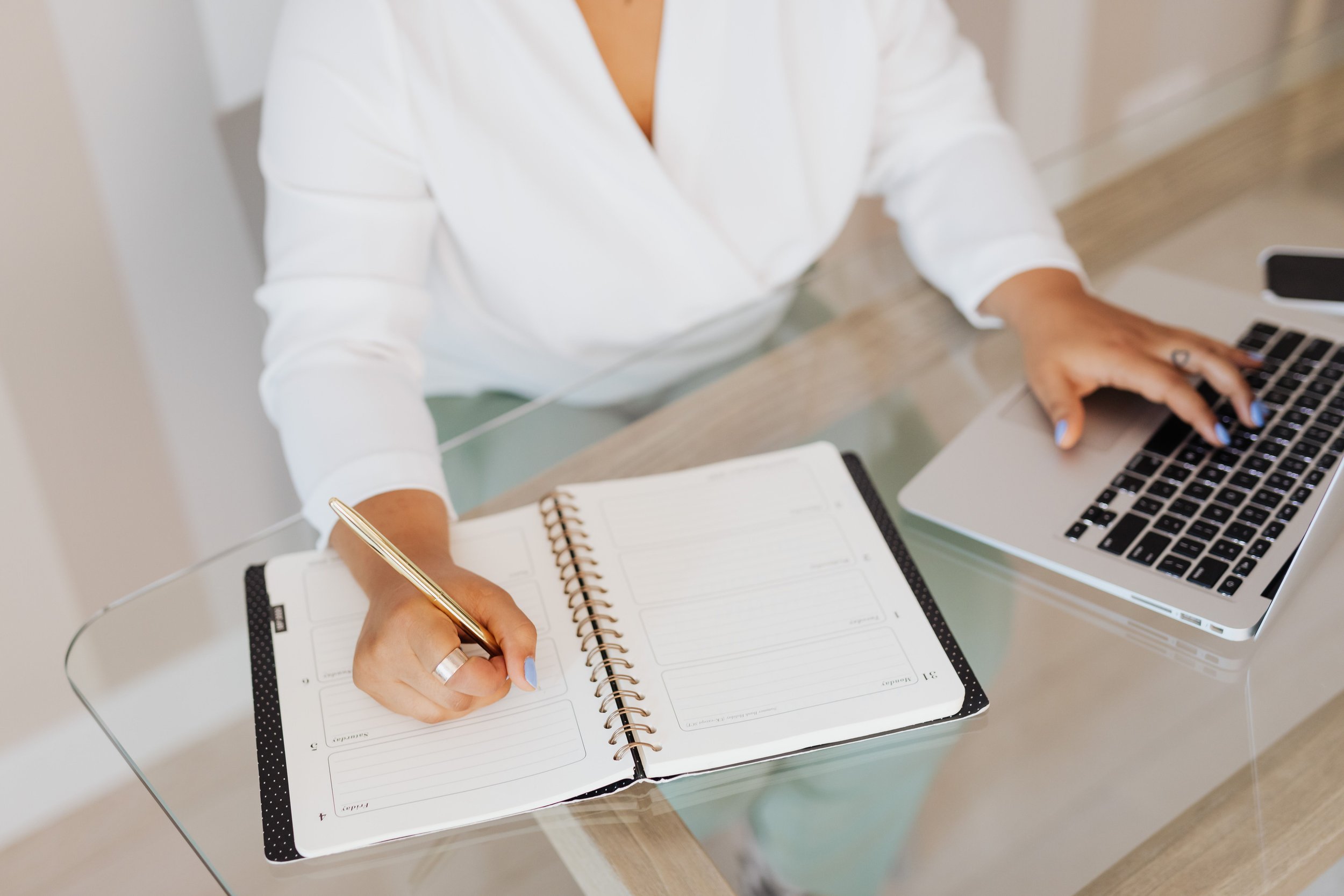 Woman in a white blazer writing in a weekly planner beside her laptop