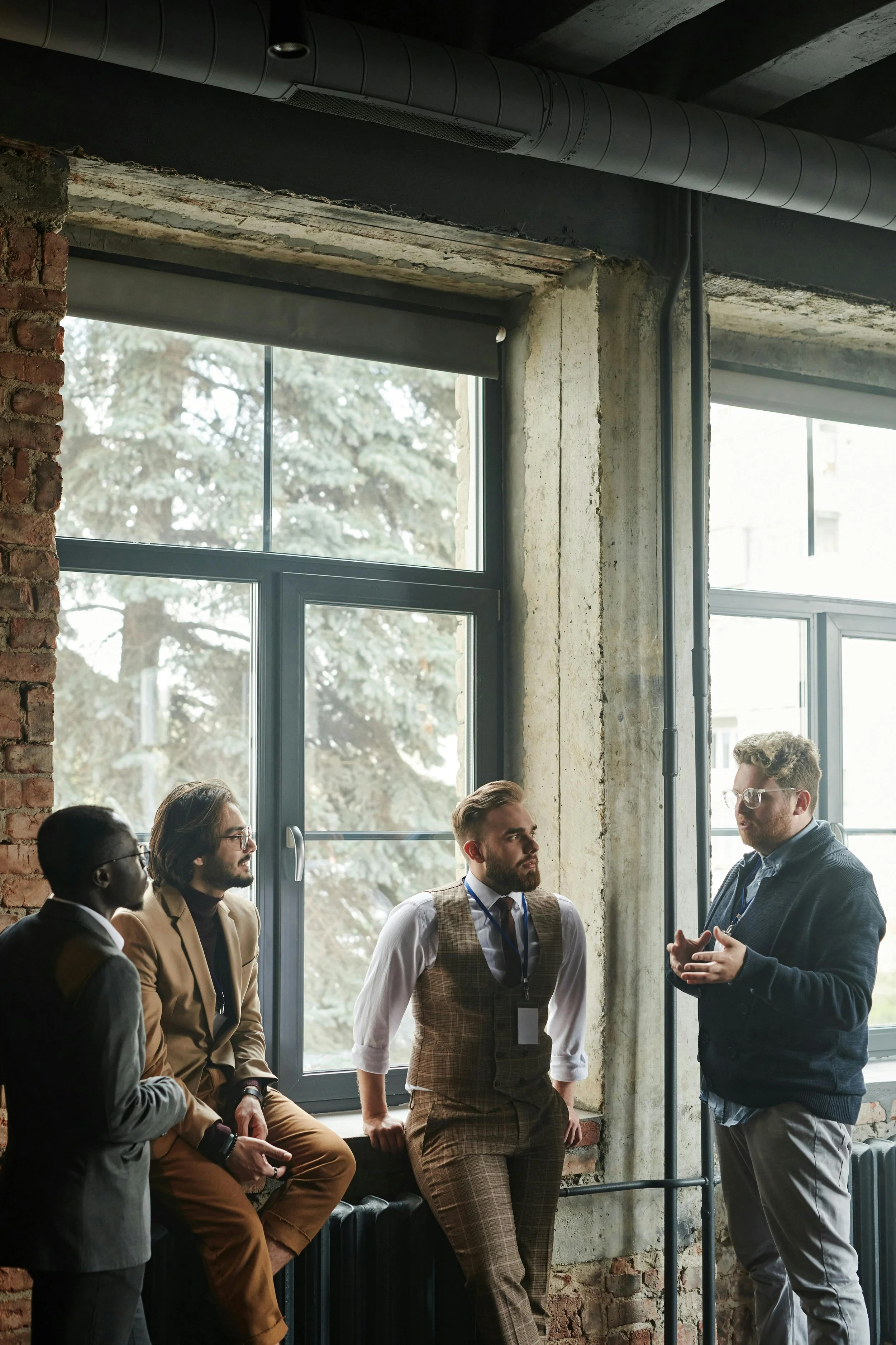 Four business professionals engaged in focused conversation in a warmly lit industrial space — representing small-group, challenge-first interactions designed to drive meaningful engagement and relationship depth.