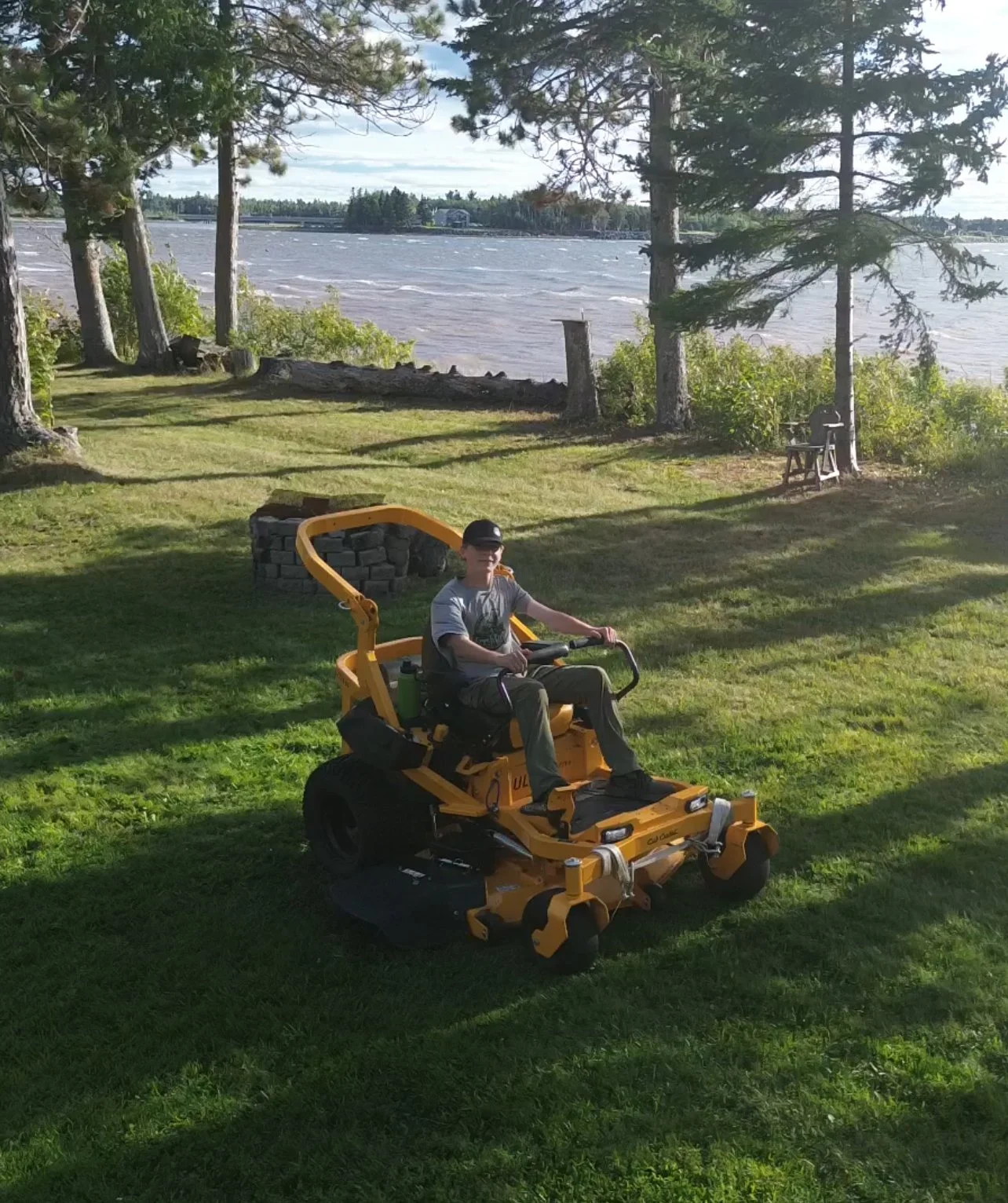 Person riding a yellow lawnmower on a grassy lawn near a lake with trees and a stack of firewood in the background.