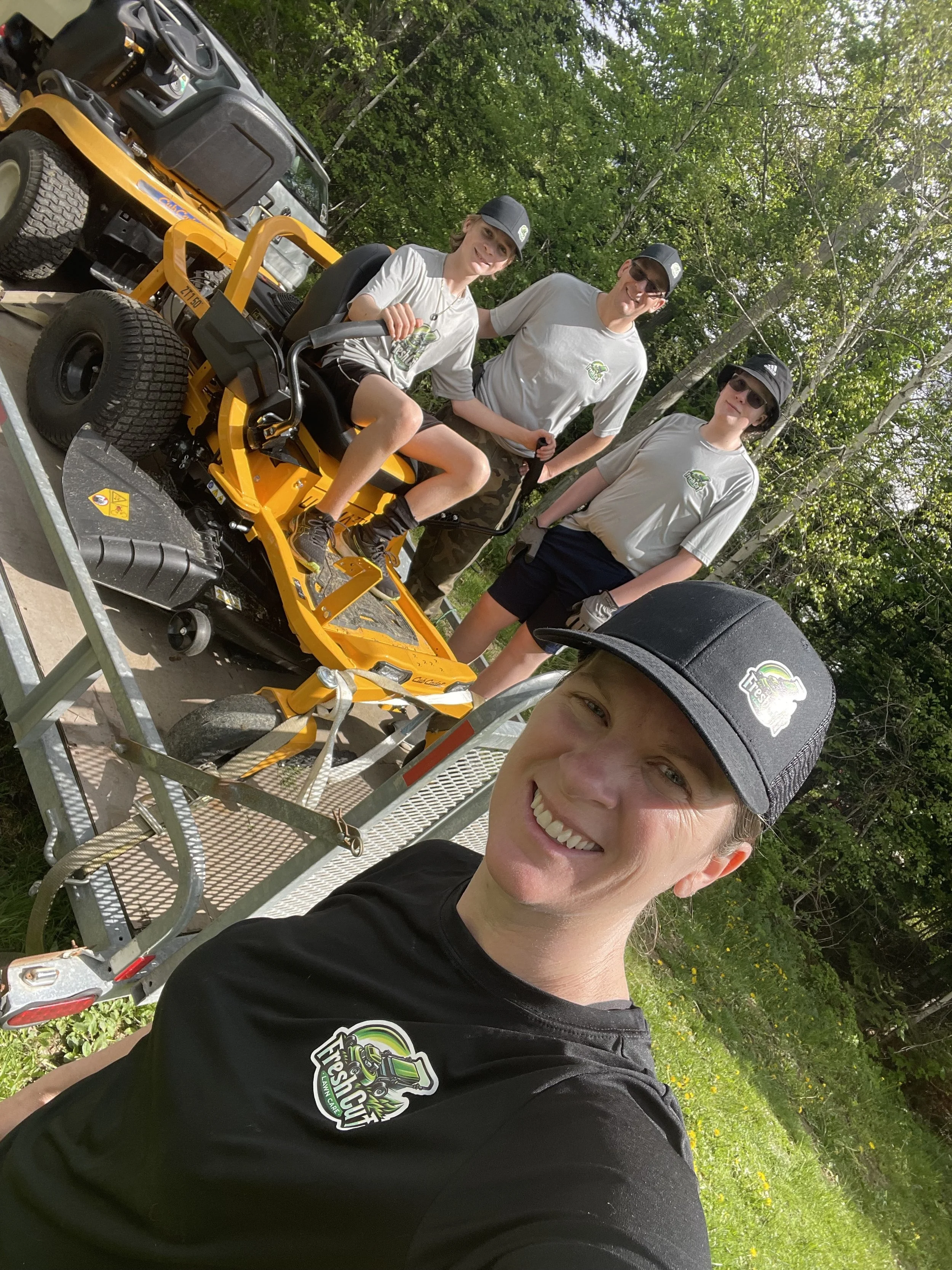 A group of four people, likely a landscaping crew, posing with a yellow riding lawn mower. They are wearing matching hats and shirts with a company logo. They are on a trailer in a grassy area surrounded by trees.