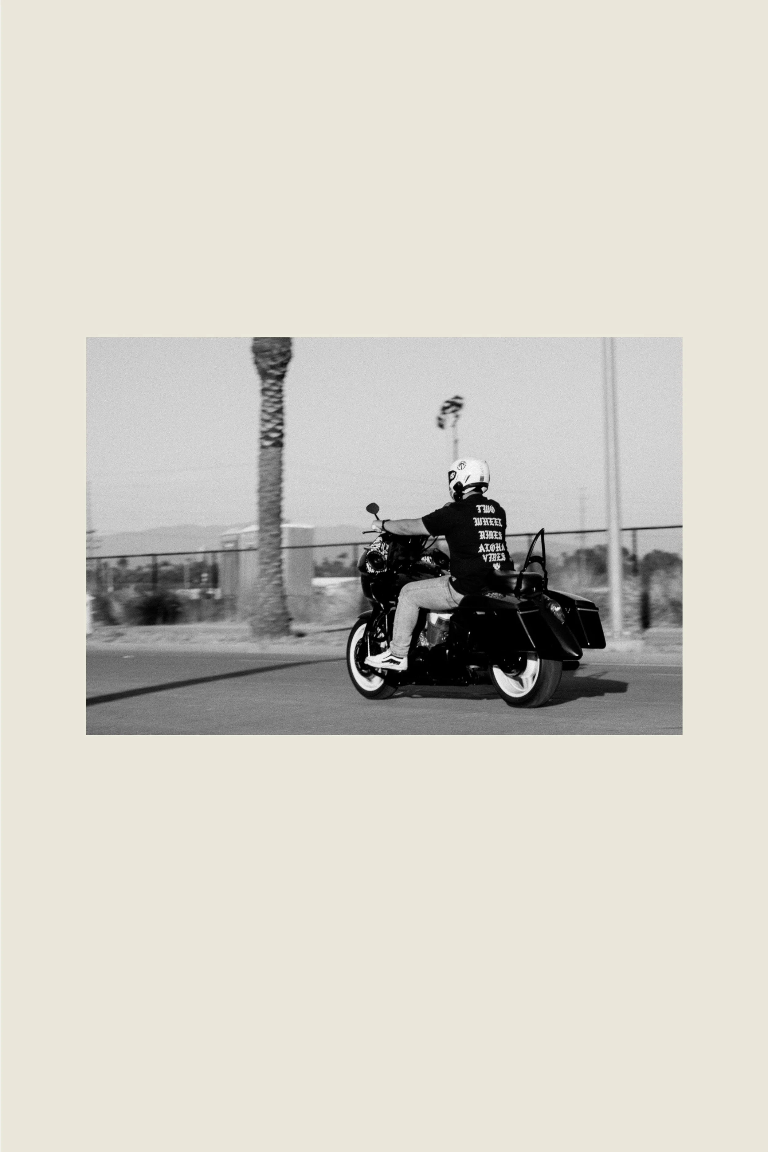 A man wearing a helmet and a dark future history T-shirt riding a black motorcycle on a street. The photo is in black and white, with palm trees and power lines in the background.