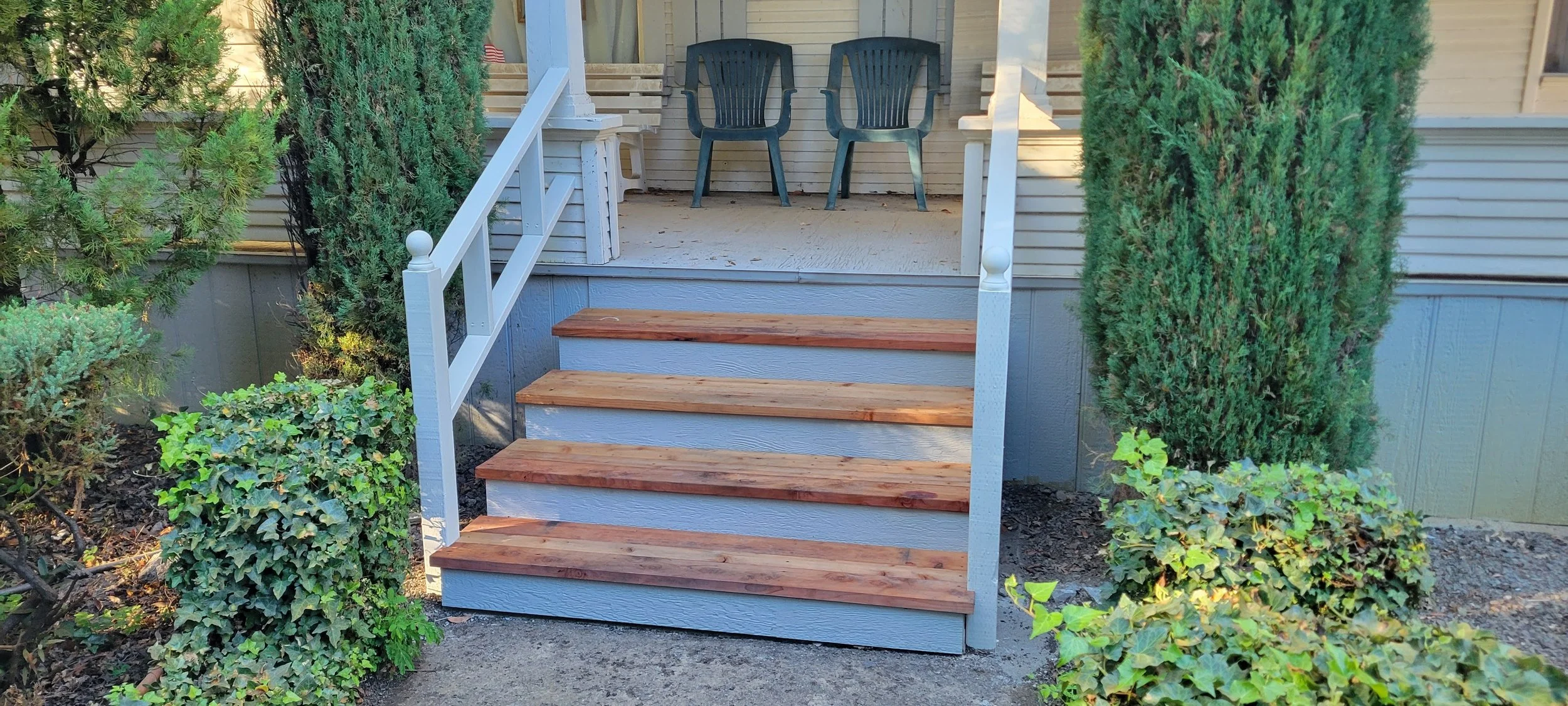 New redwood front porch steps with brand new white railings. The porch has a white wooden bench. Surrounding the steps are green shrubs and trees.
