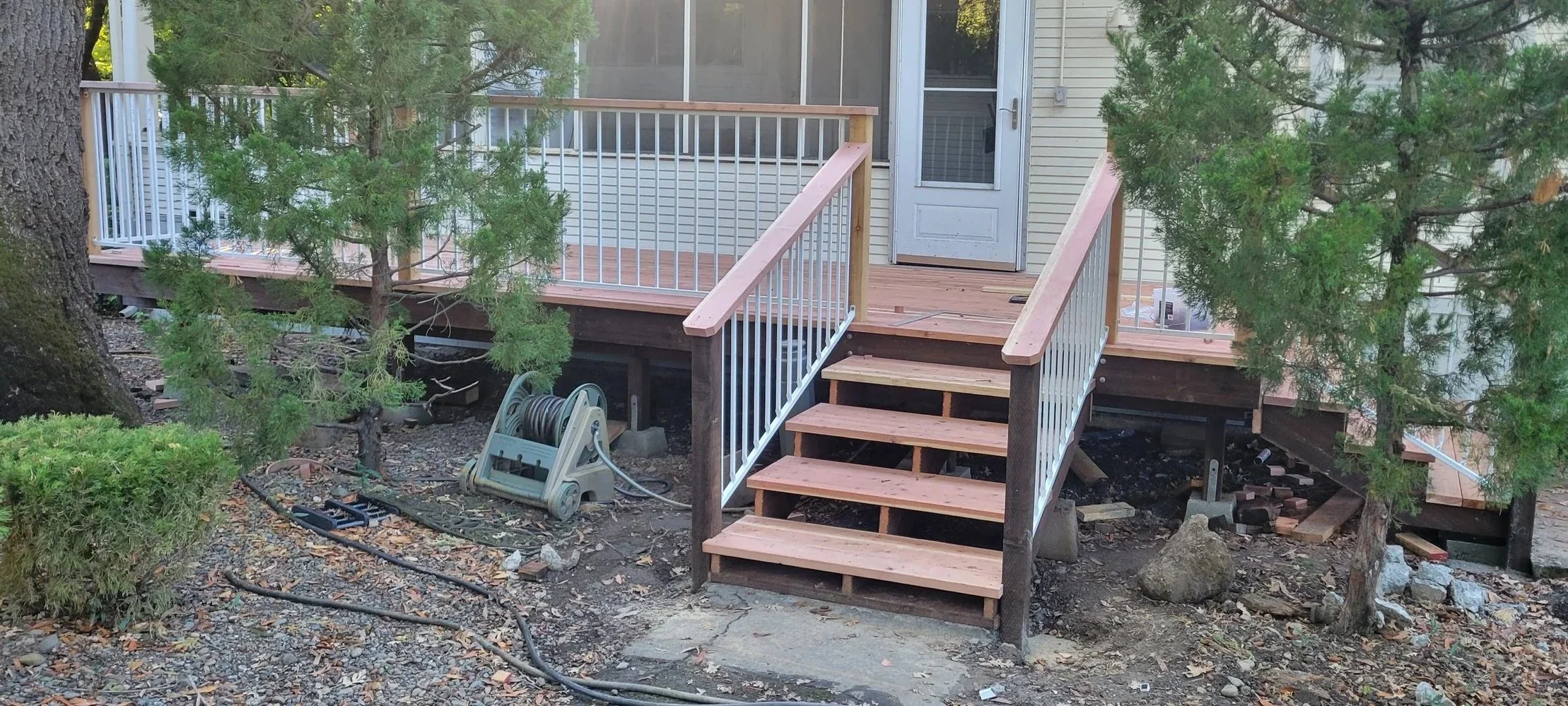 New beautiful redwood deck with stairs and clean white powder coated steele railing finished construction, surrounded by trees, tools, and construction materials on the ground.