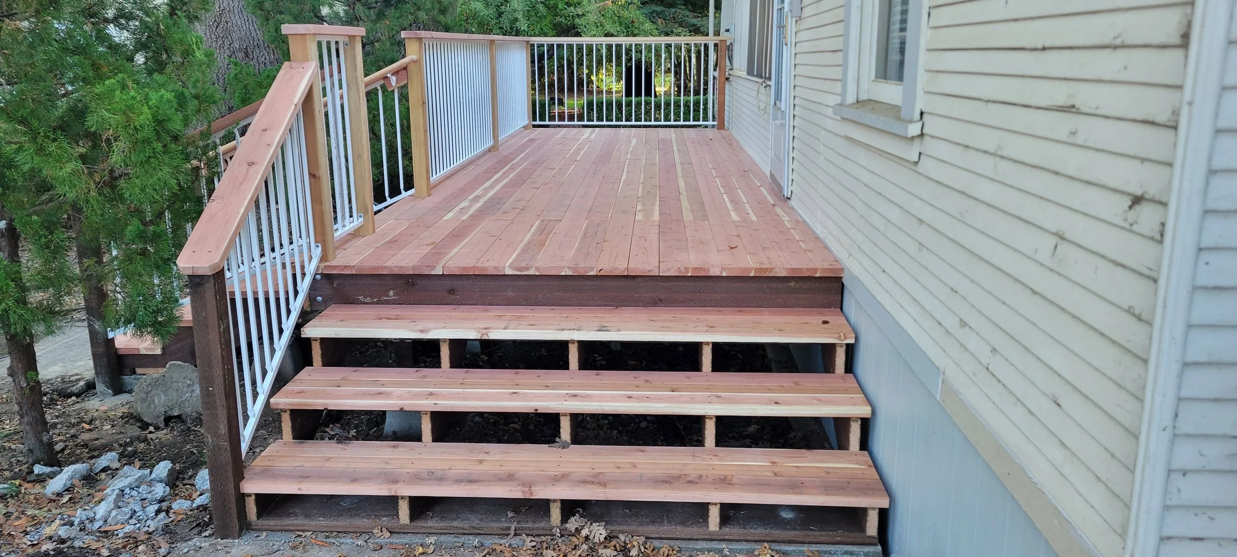 A newly constructed wooden deck attached to a house, with stairs leading down to the ground, enclosed by a metal and wood railing, with a background of green trees and yard.