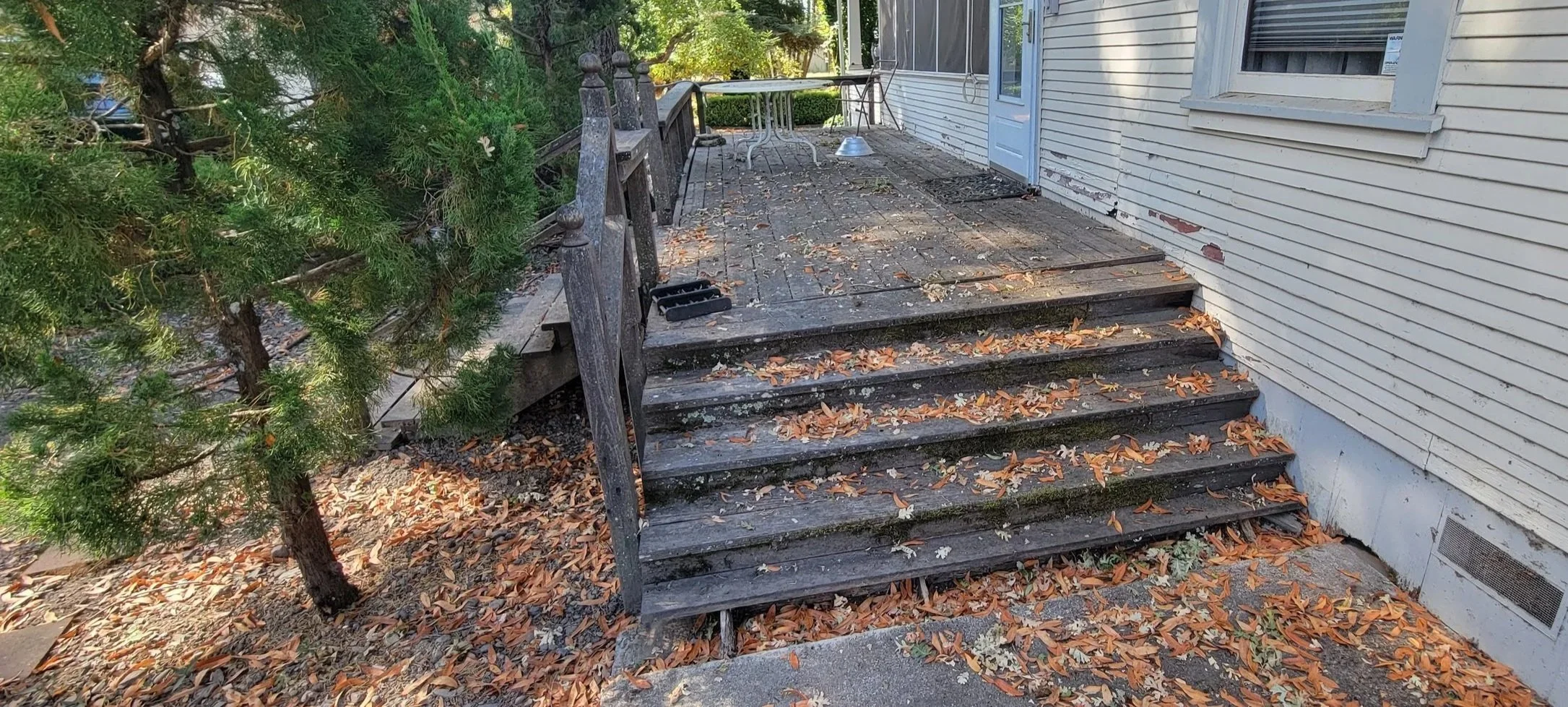 Back porch with worn wooden stairs, leaf-covered ground, a house with white siding, a door, and a window. There is a tree on the left and some outdoor furniture on the porch.