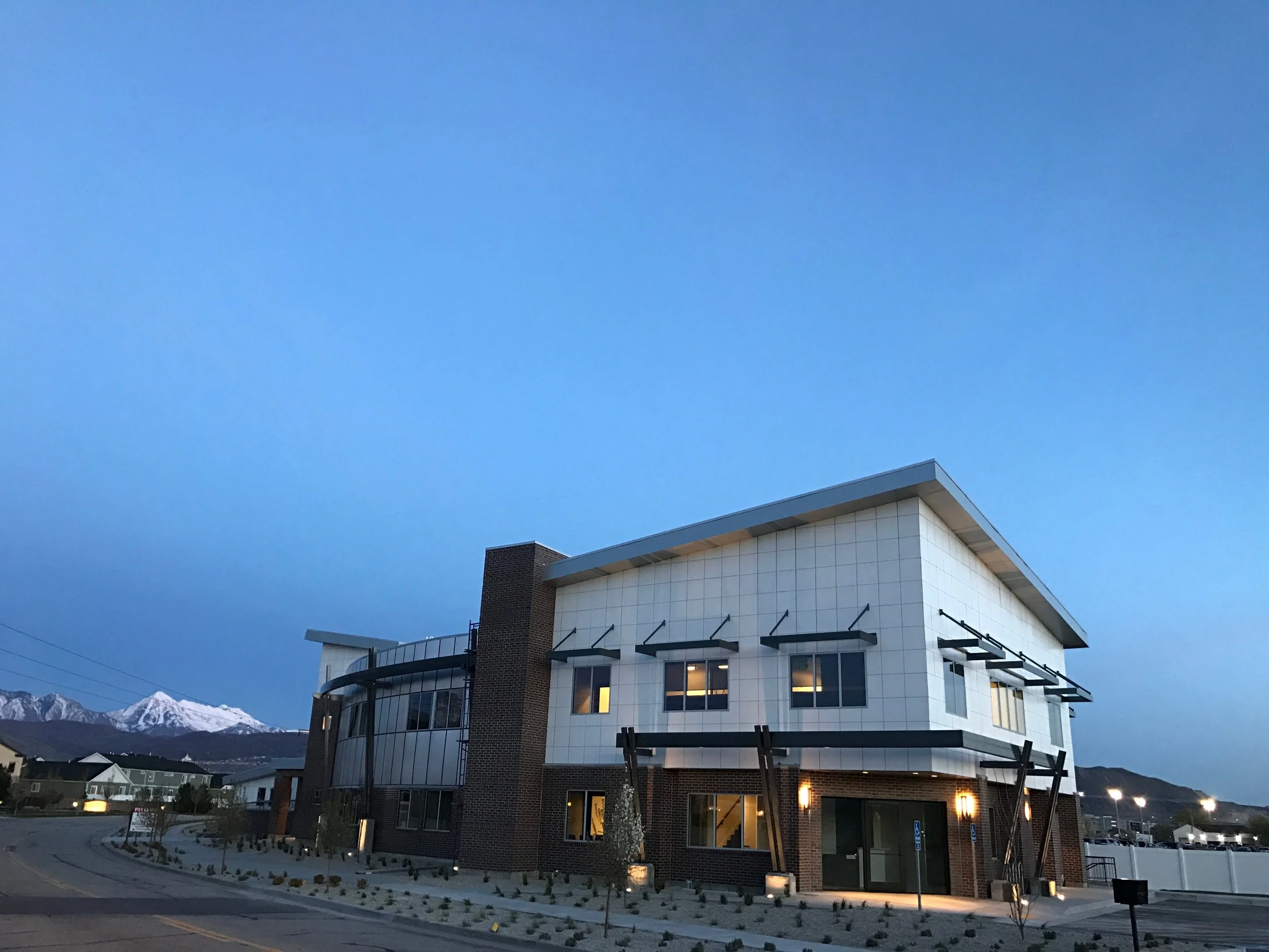 A modern two-story building with brick and white panel exterior, lit windows, and surrounding landscape, with snow-capped mountains in the background during dusk.