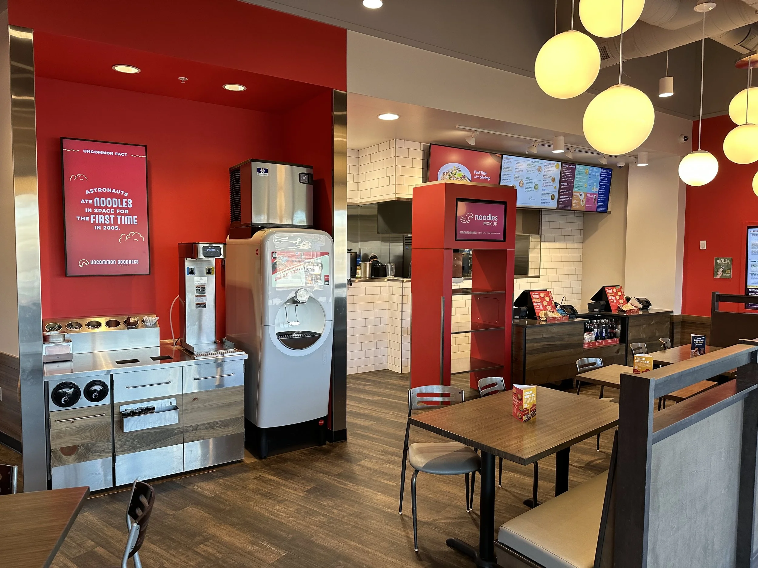 Inside a fast-casual restaurant with red and white walls, tables, chairs, digital menu screens, and a self-serve frozen yogurt station.