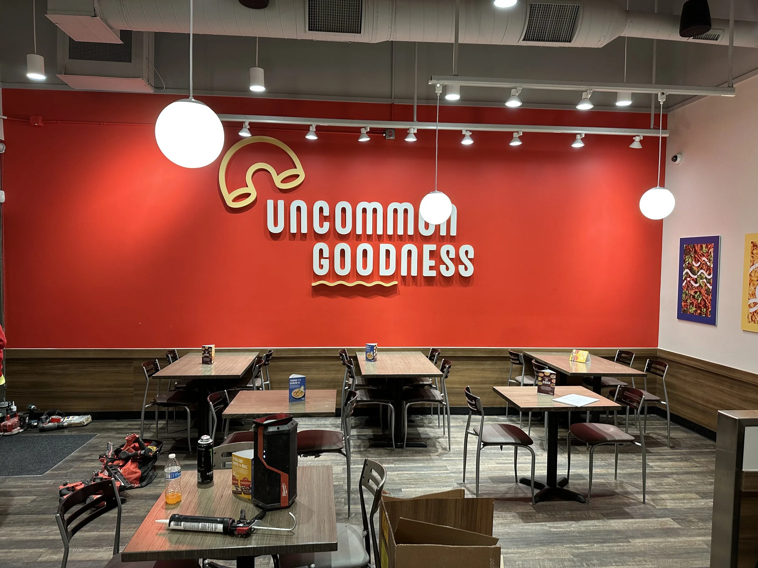 Empty fast food restaurant with wooden tables and chairs, bright red wall with a logo and text 'Uncommon Goodness', and colorful artwork on the sides.