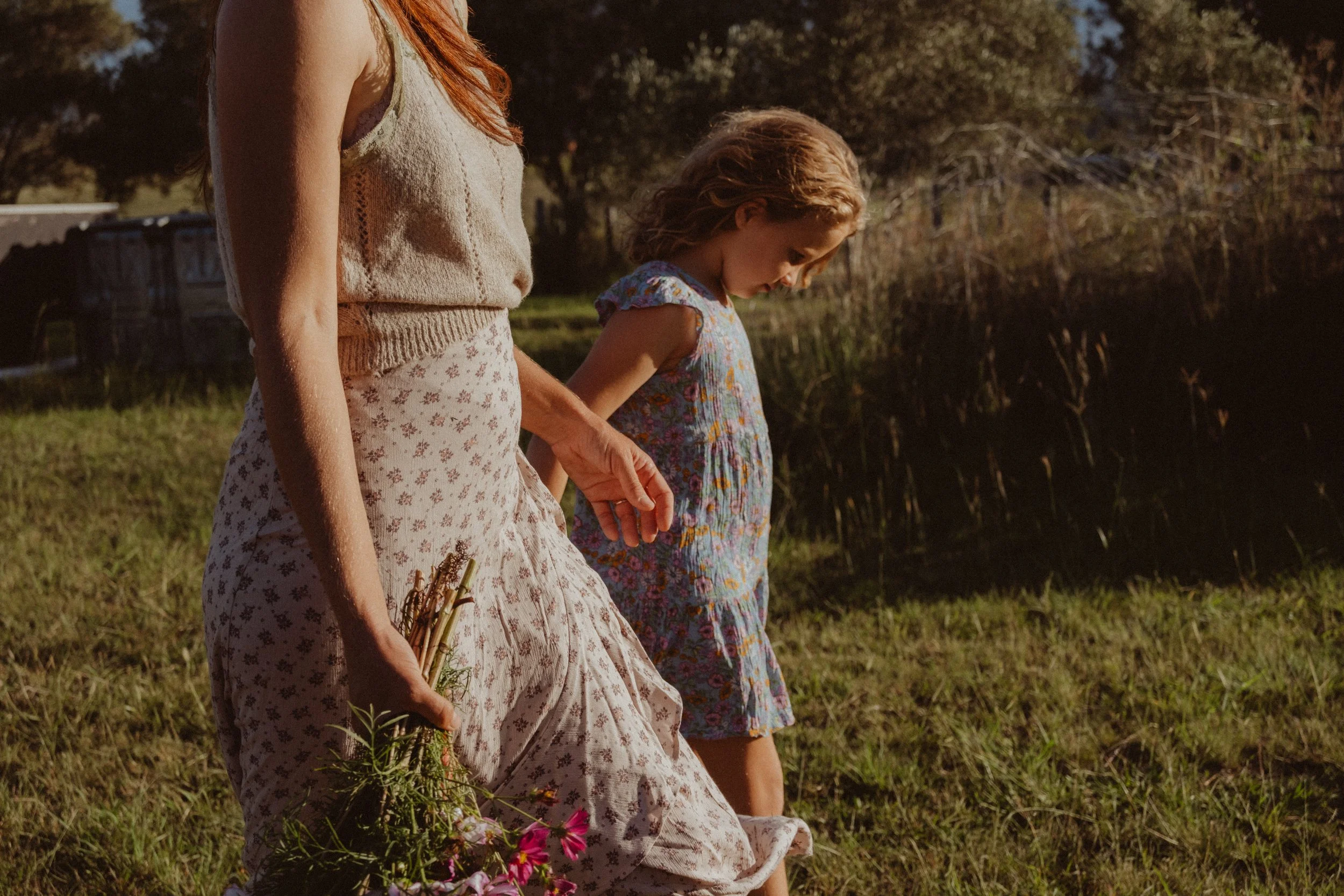 A woman and a young girl walking outdoors on a grassy field in the late afternoon or early evening, with trees and a house visible in the background.