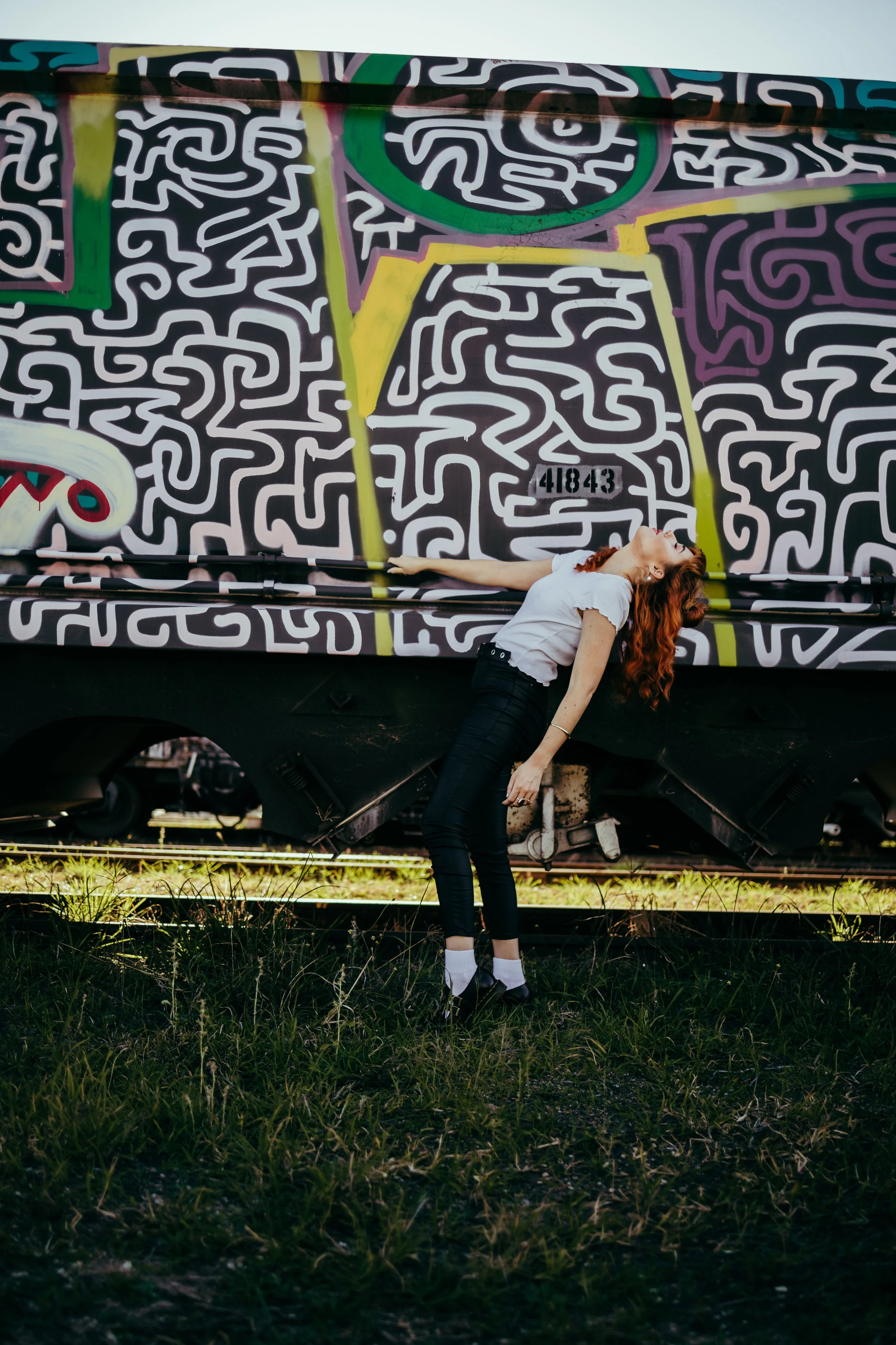 A young woman with red hair leaning backward with arms extended, standing in front of a graffitied train car on a grassy area.