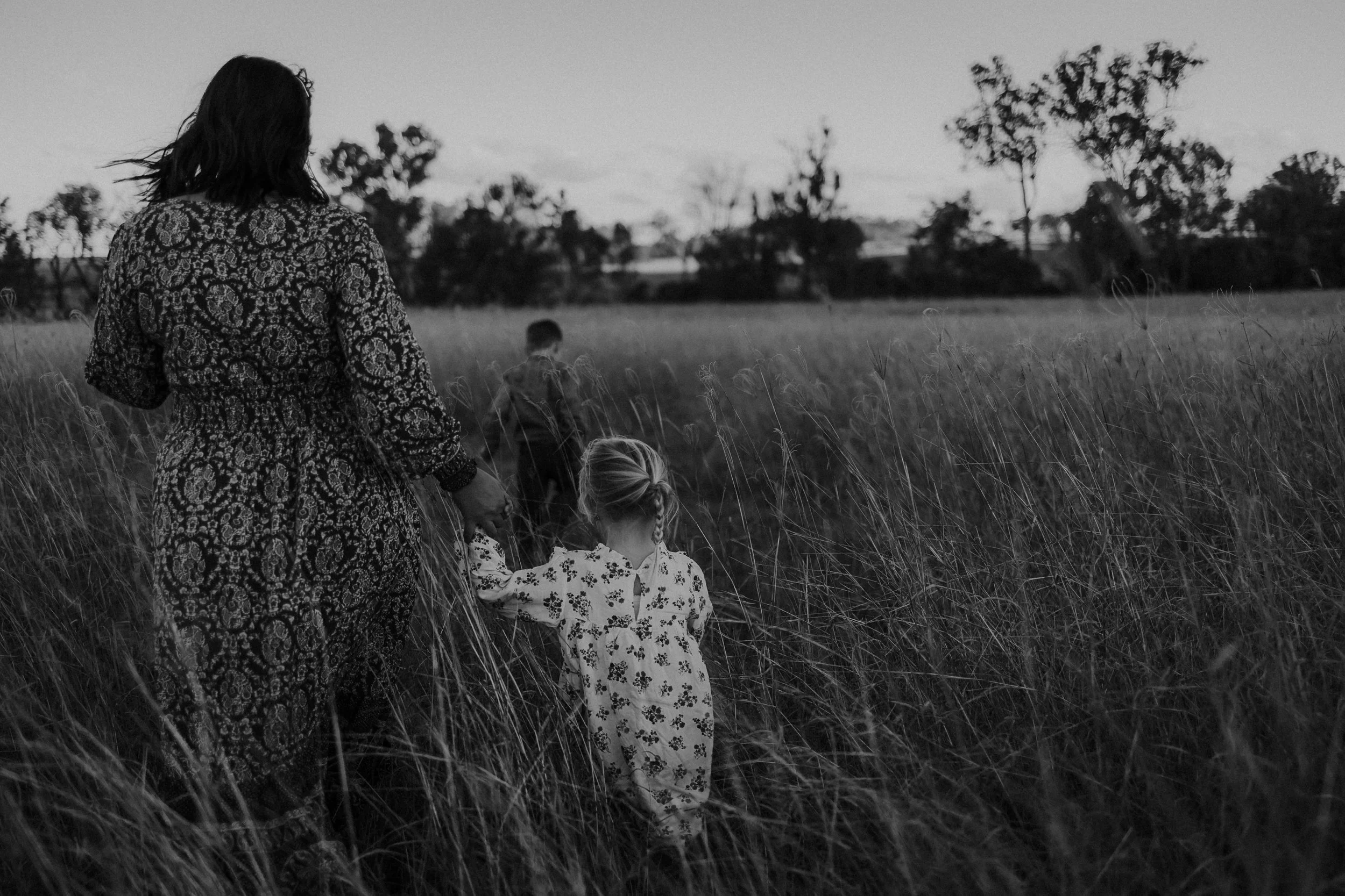 A woman and two children walking through a grassy field, holding hands, with trees in the background, black and white photograph.