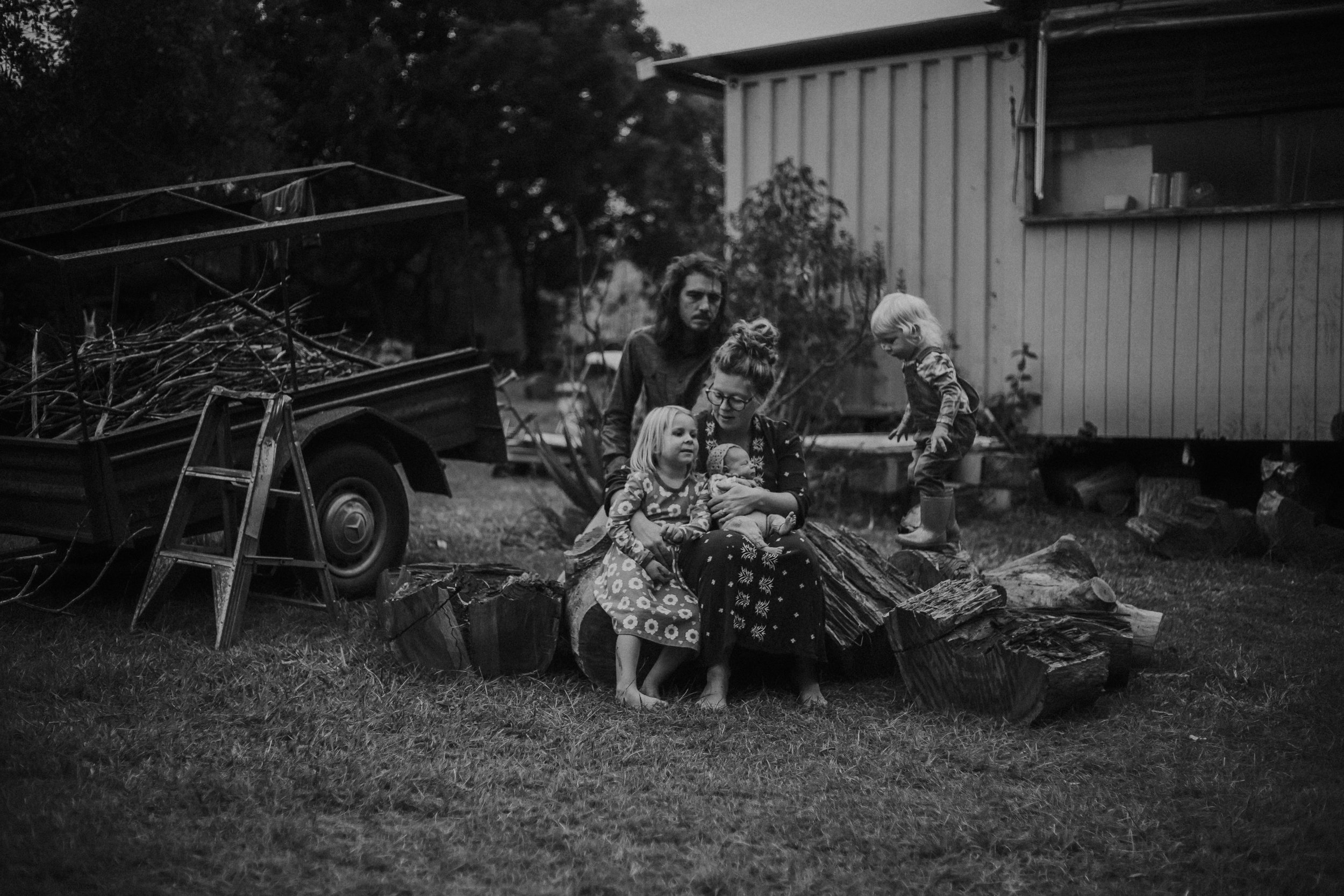 A black and white photo of a family sitting outdoors surrounded by logs and wood pieces, with children playing nearby.