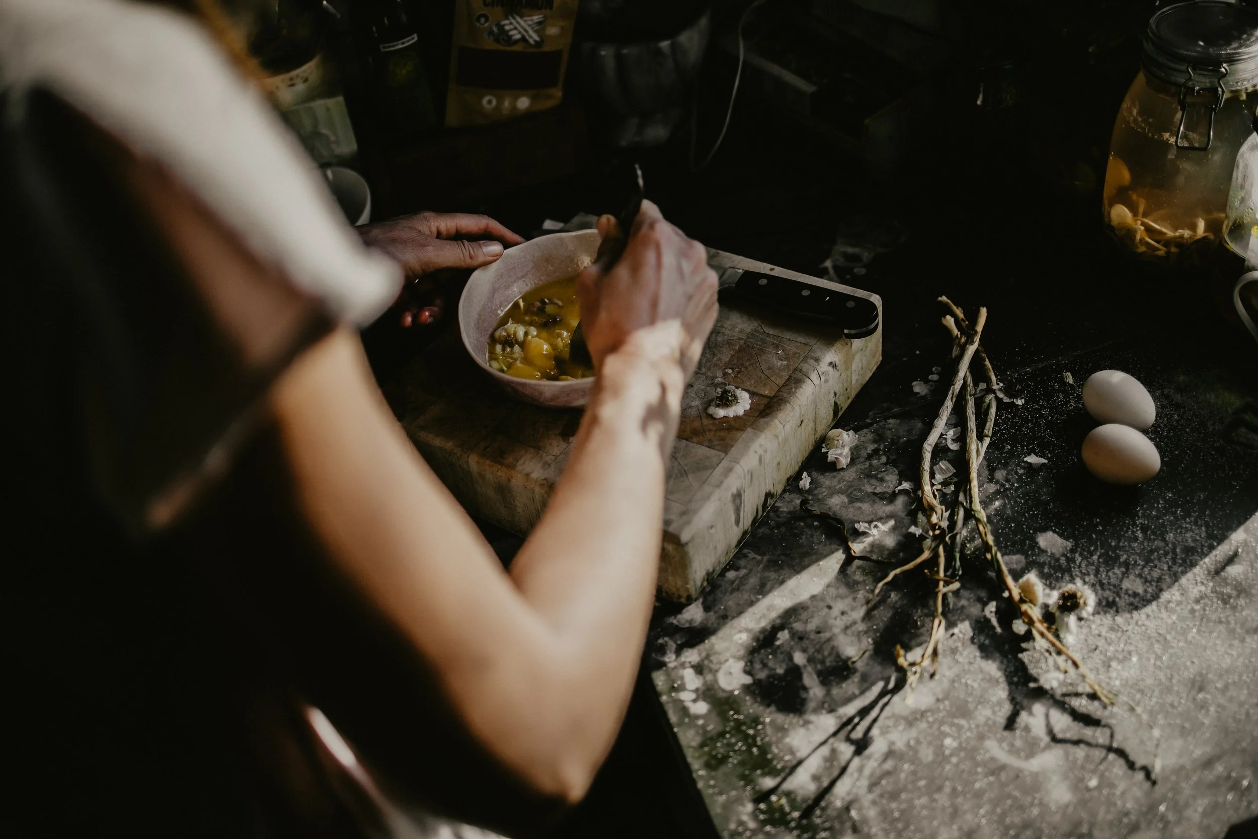 A person mixing ingredients in a pink bowl on a kitchen counter, with eggs, dried flowers, and kitchen supplies around.