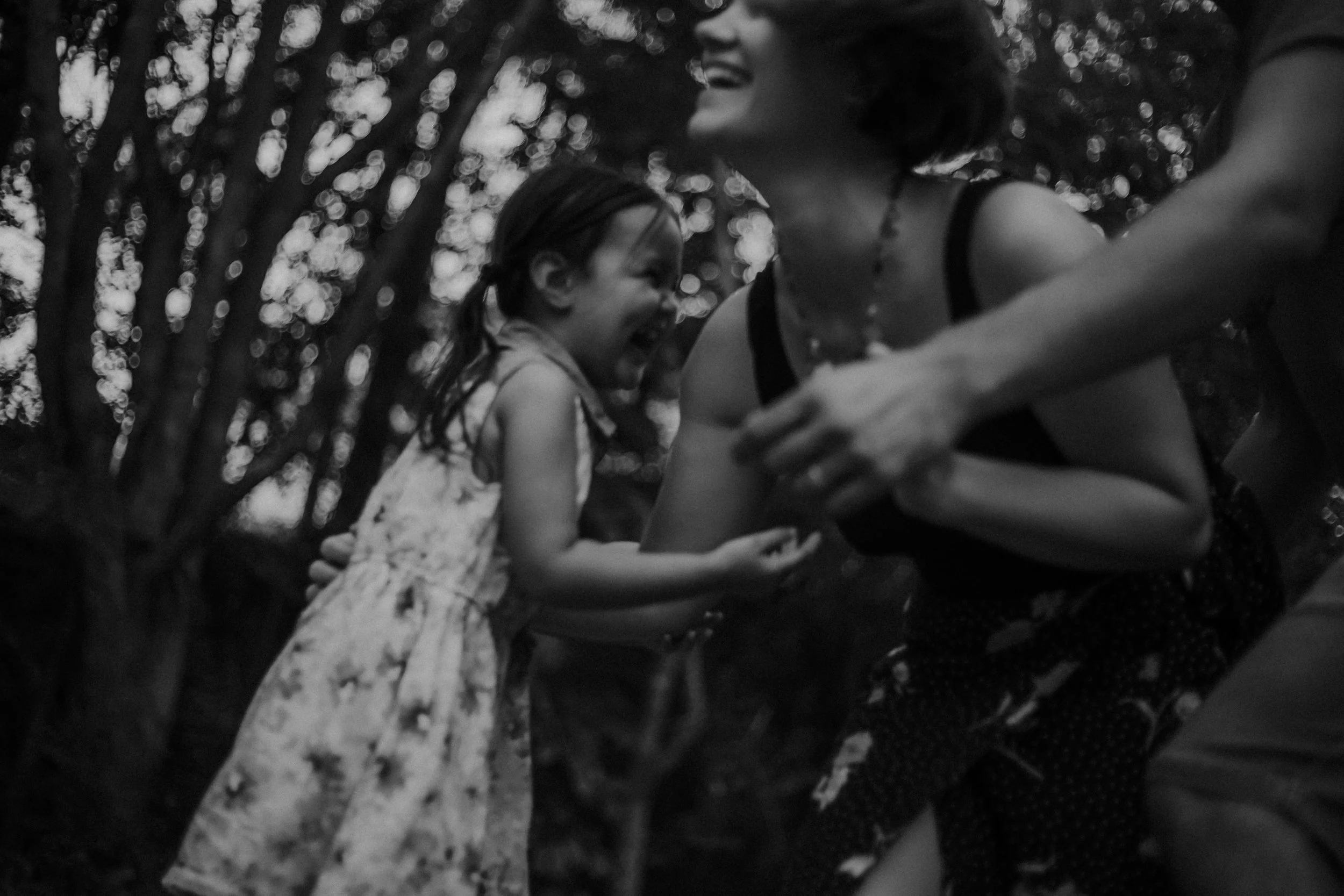 A black and white photo of a woman laughing and a young girl smiling, with another person's arm in the foreground, outdoors with trees in the background.