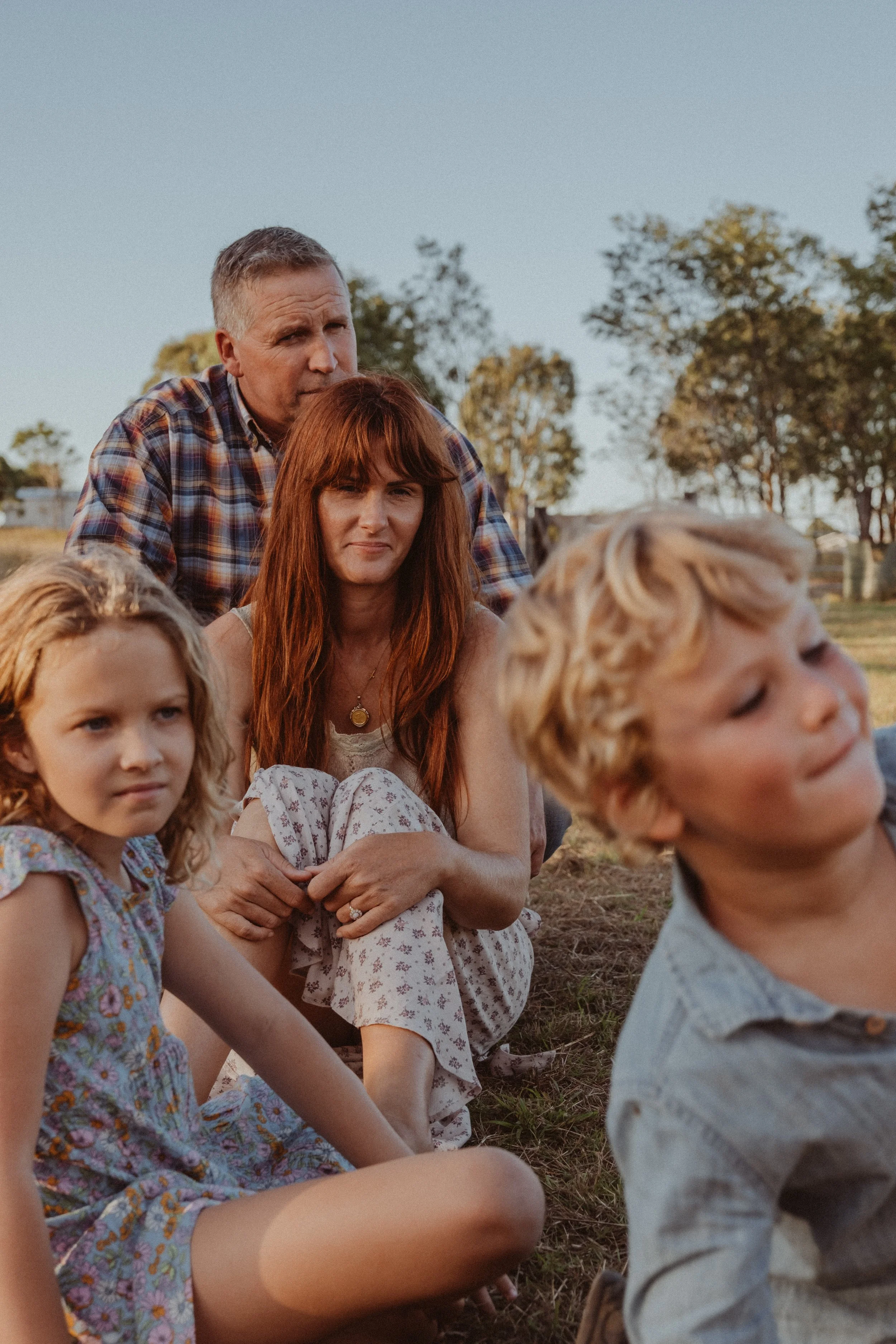 A family sitting outdoors on grass in a park during late afternoon, with trees and a clear sky in the background.