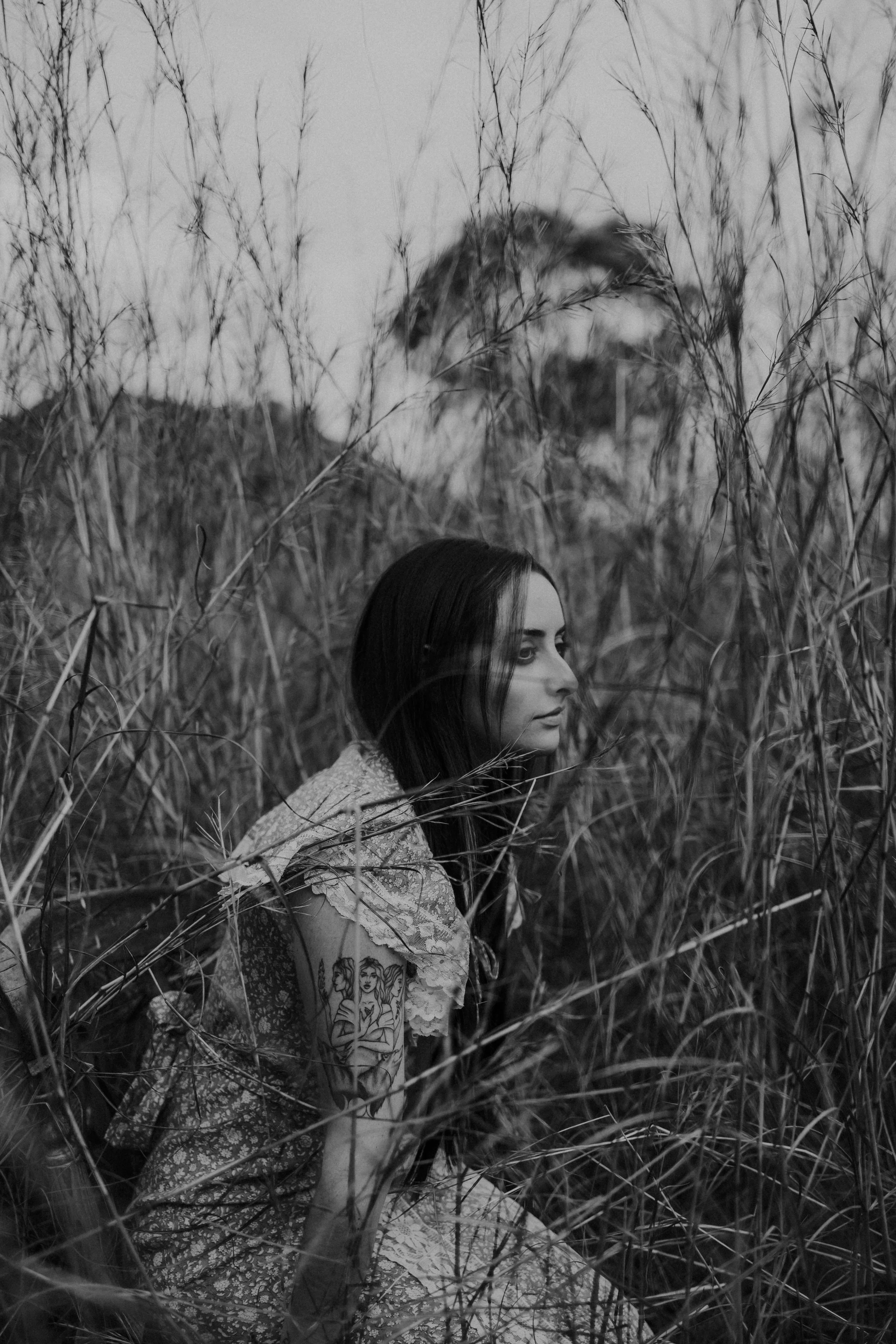A woman with long dark hair and tattoos on her arm, sitting in tall grass, with a faint image of her face overlaid in the background.