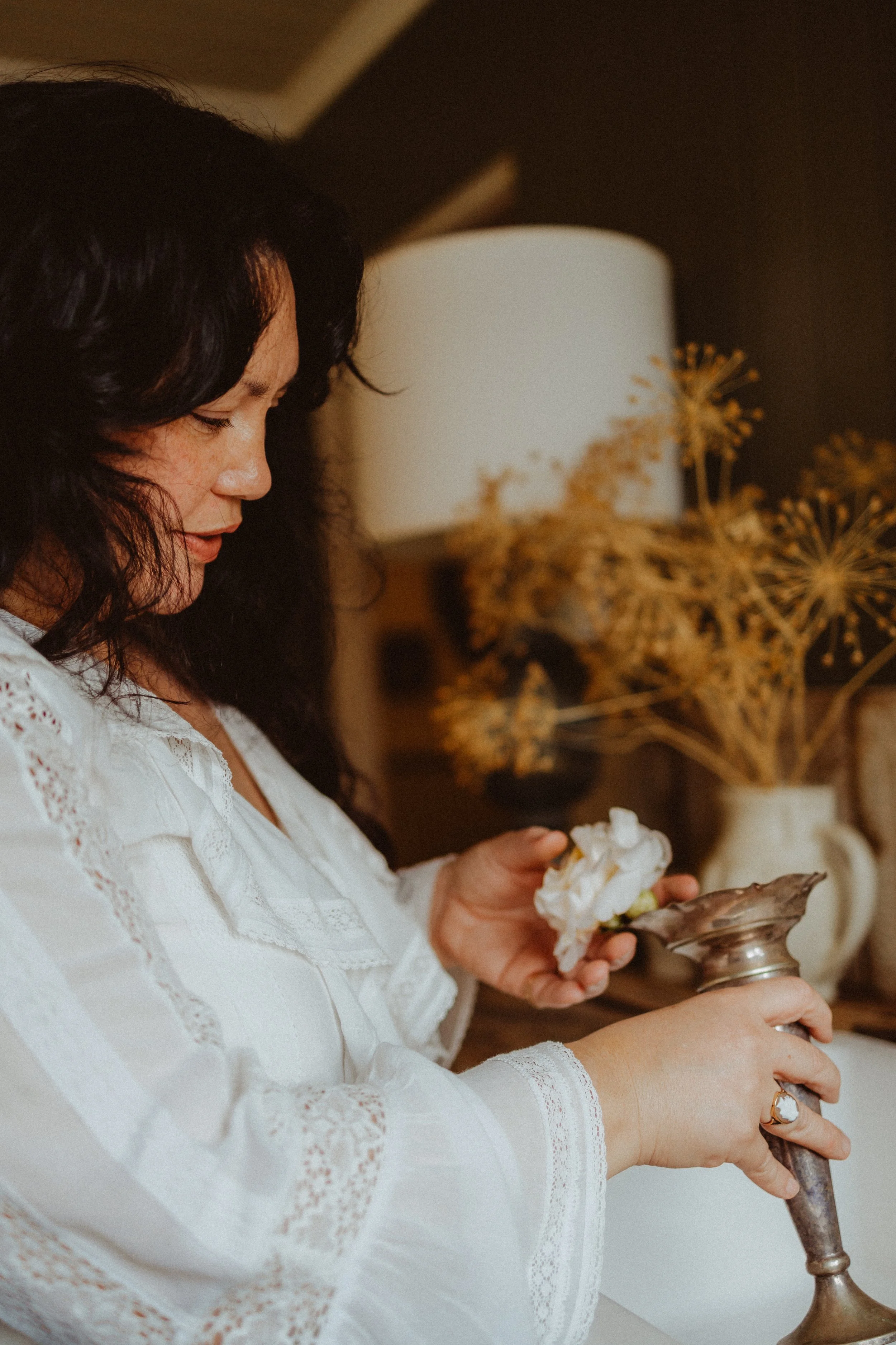 A woman with dark, wavy hair wearing a white lace blouse holding a flower and a metal object, with a lamp and dried plants in the background.