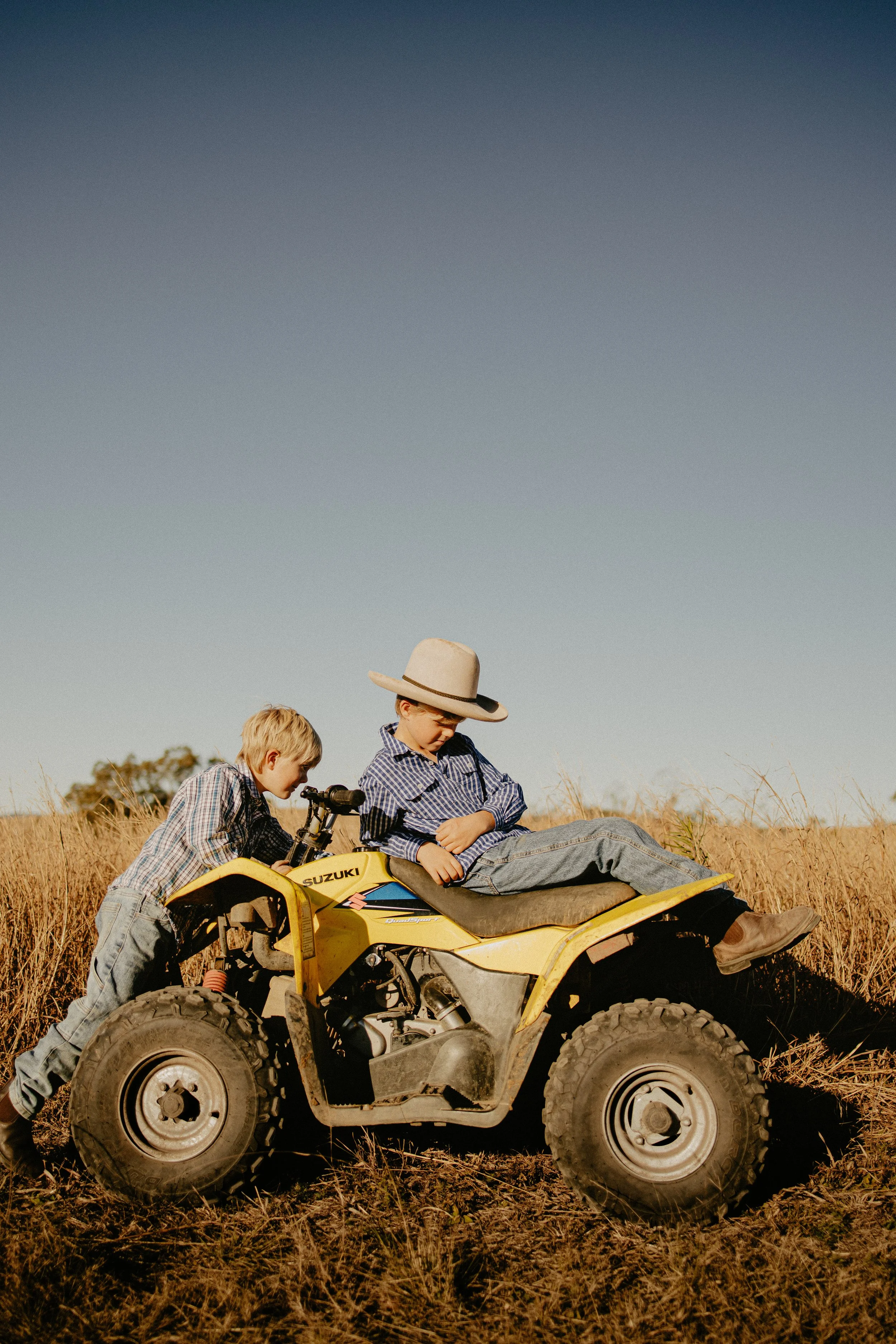 Two young boys in cowboy hats and plaid shirts with jeans, one sitting on a yellow Suzuki all-terrain vehicle (ATV) and the other leaning on it in a field of tall grass under a clear blue sky.