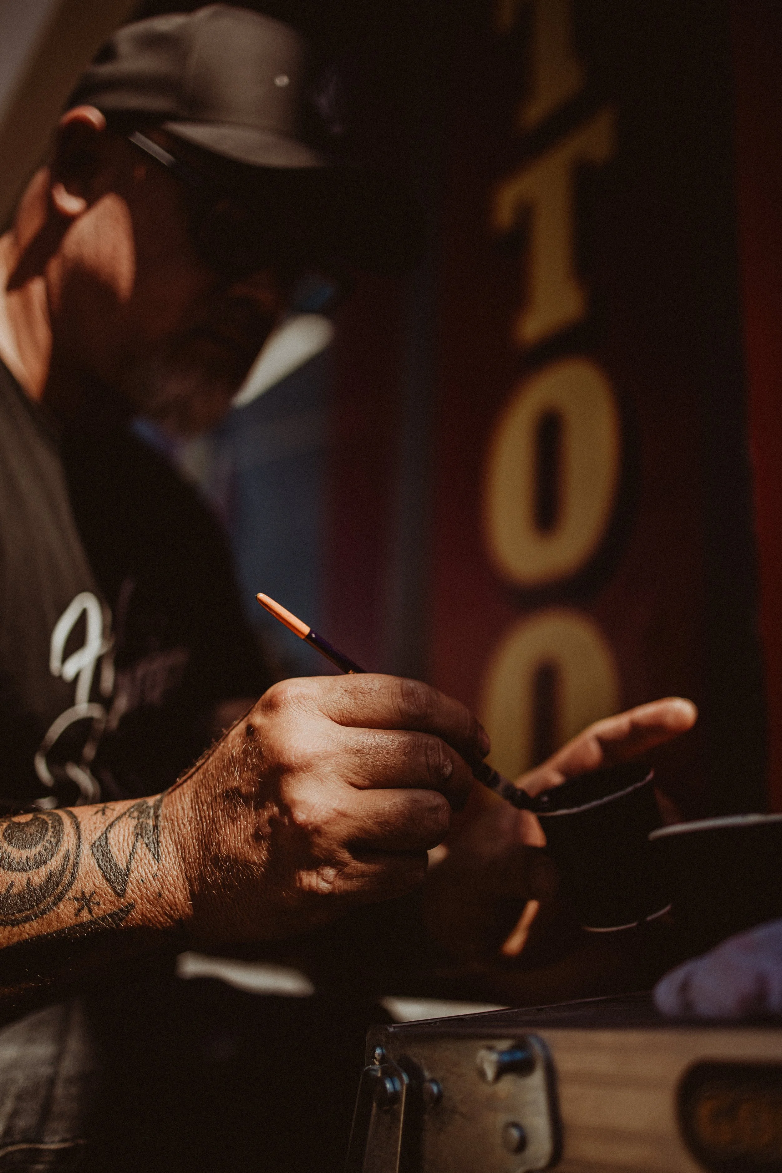 A tattooed man wearing a cap and sunglasses writes on a notepad with a pencil in a dimly lit setting, with a large, partially visible word on the background wall.