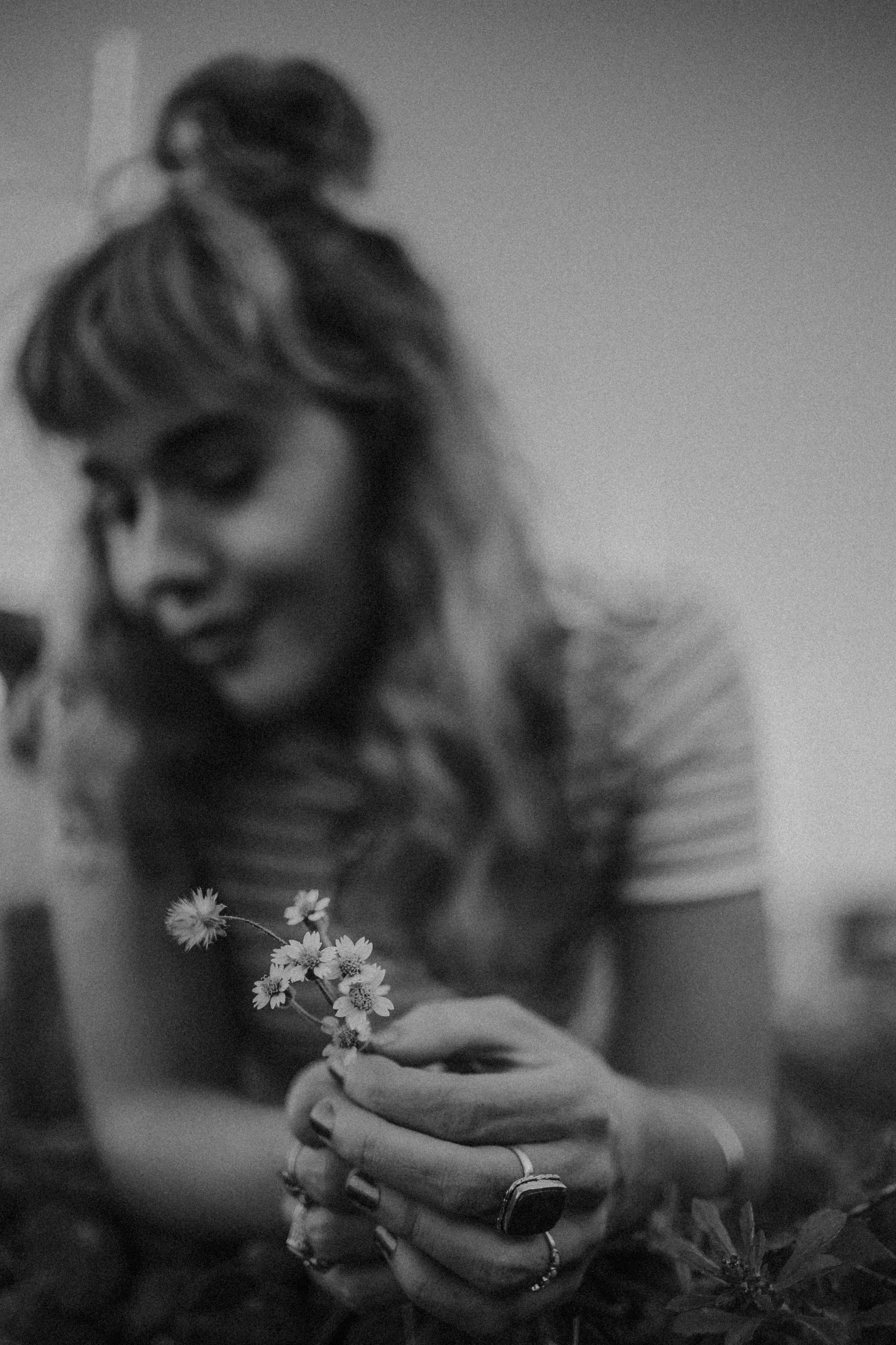 A woman with long hair, wearing rings and jewelry, holding a small bouquet of flowers, with a blurred background.