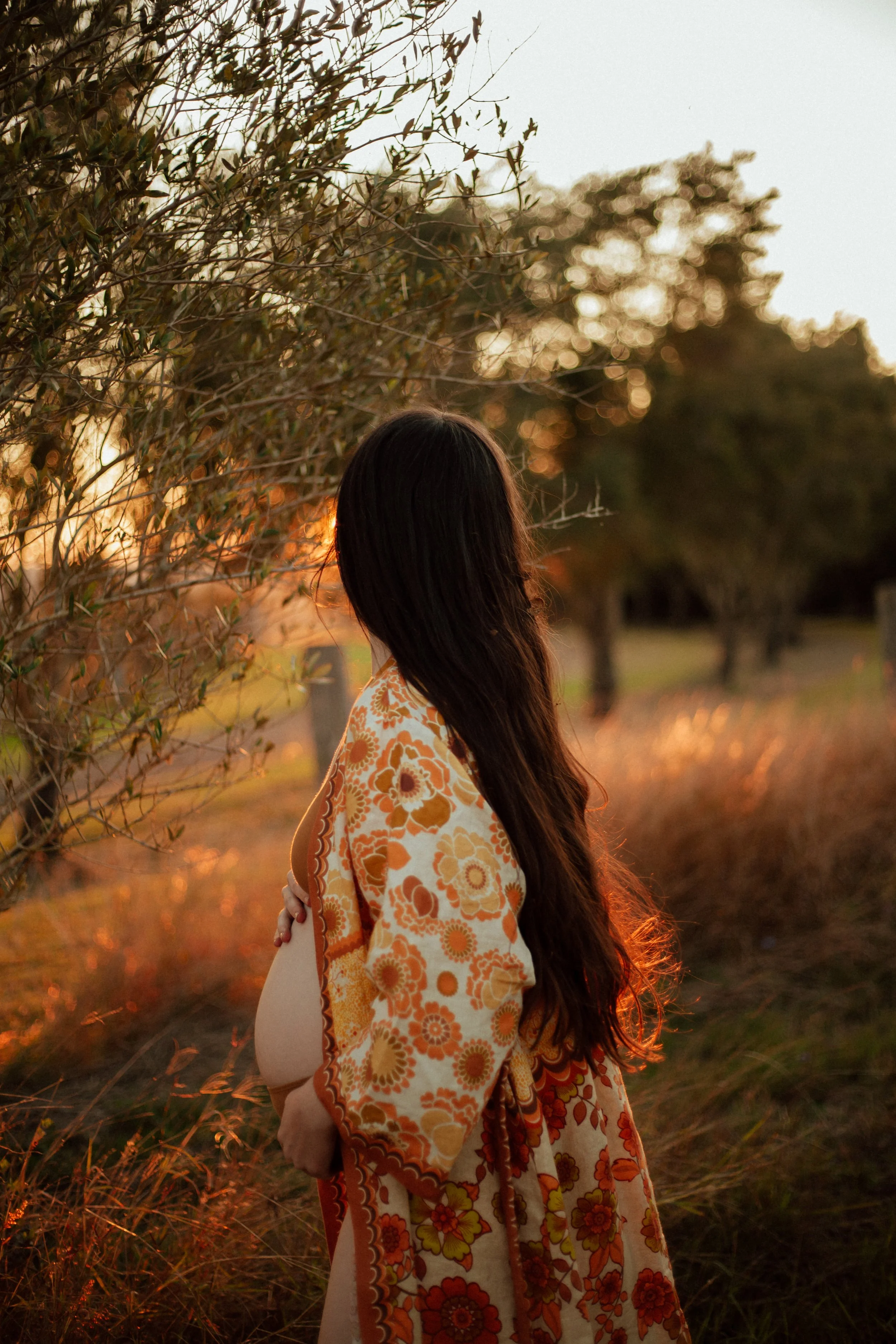 A pregnant woman with long dark hair stands outdoors during sunset, wearing a floral patterned dress, with her face turned away from the camera, touching her belly.